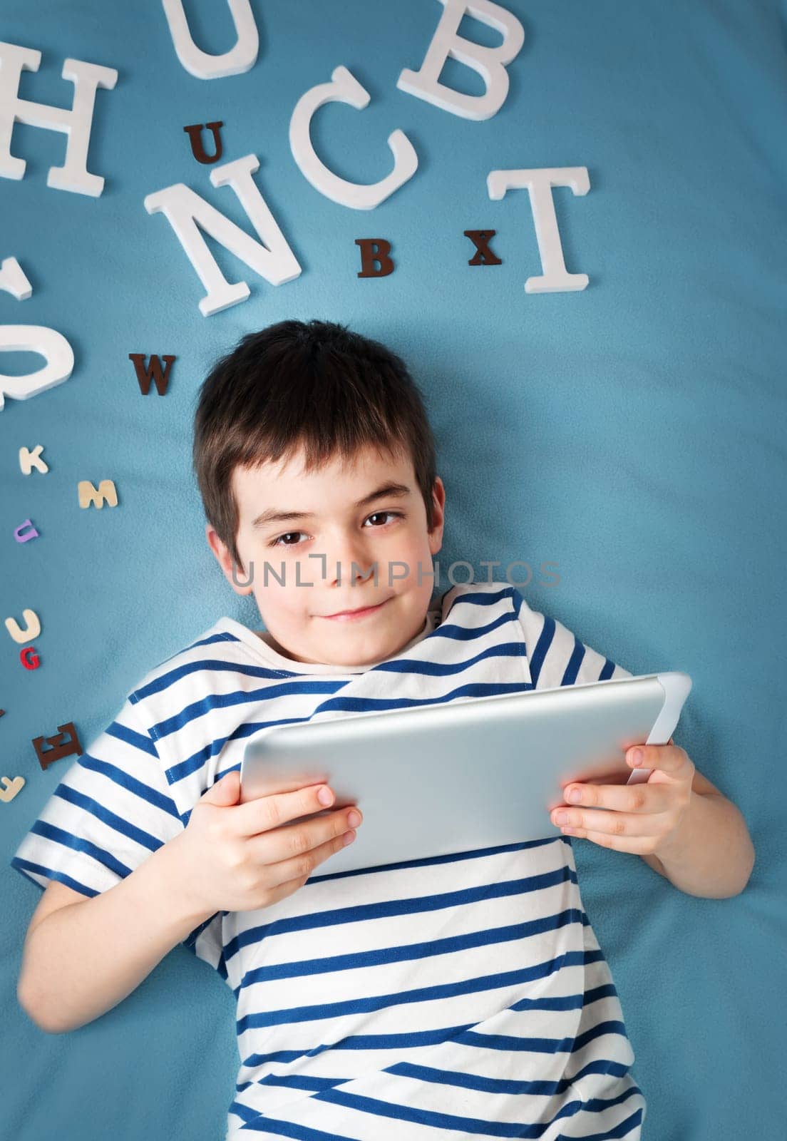 seven years old child lying with tablet and letters on blue background