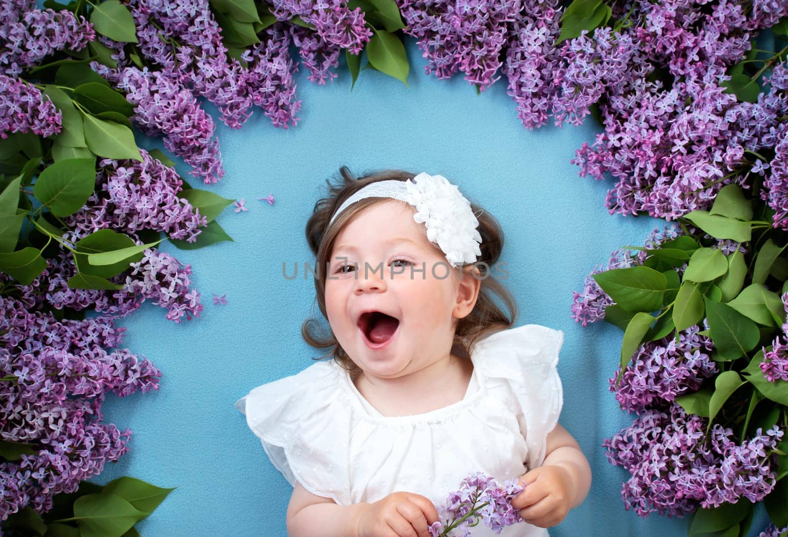 pretty little baby girl lying on blue background with lilac flowers