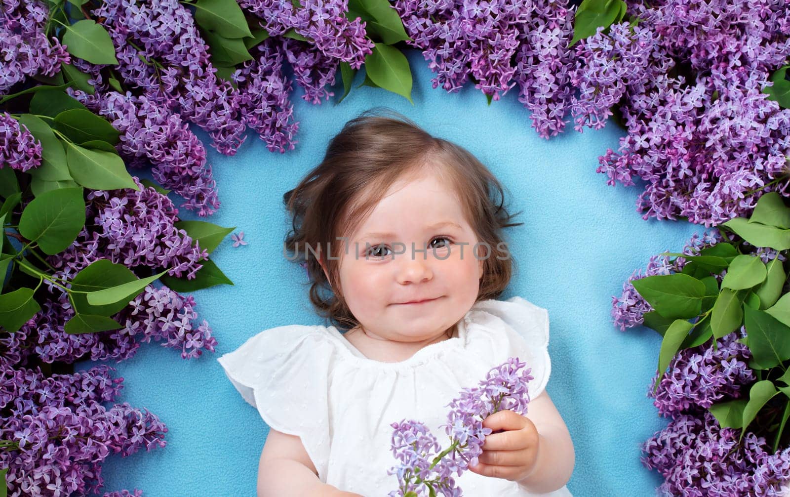 pretty little baby girl lying on blue background with lilac flowers