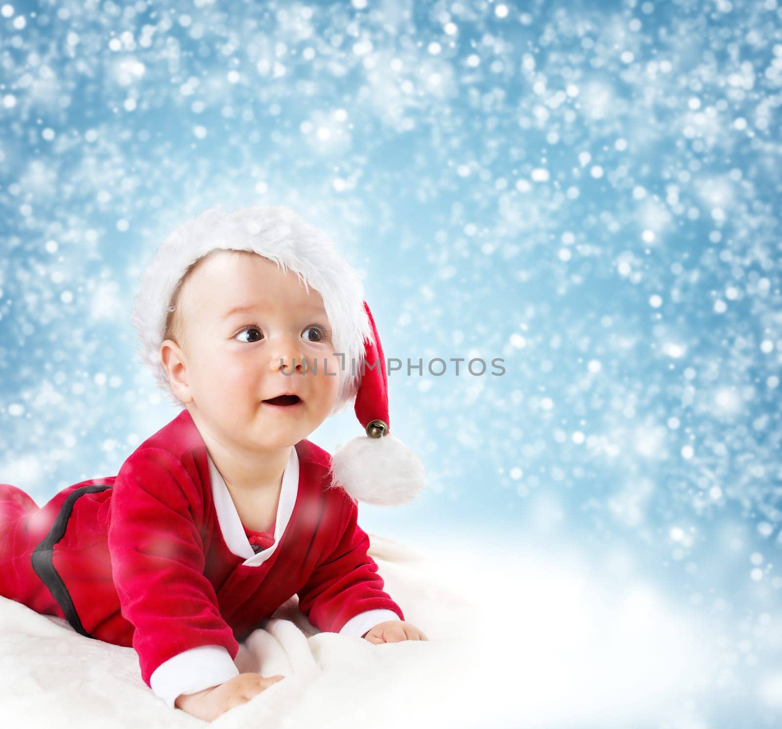 Eight month old baby in knitted winter hat lying on soft blanket with blue background and falling snow