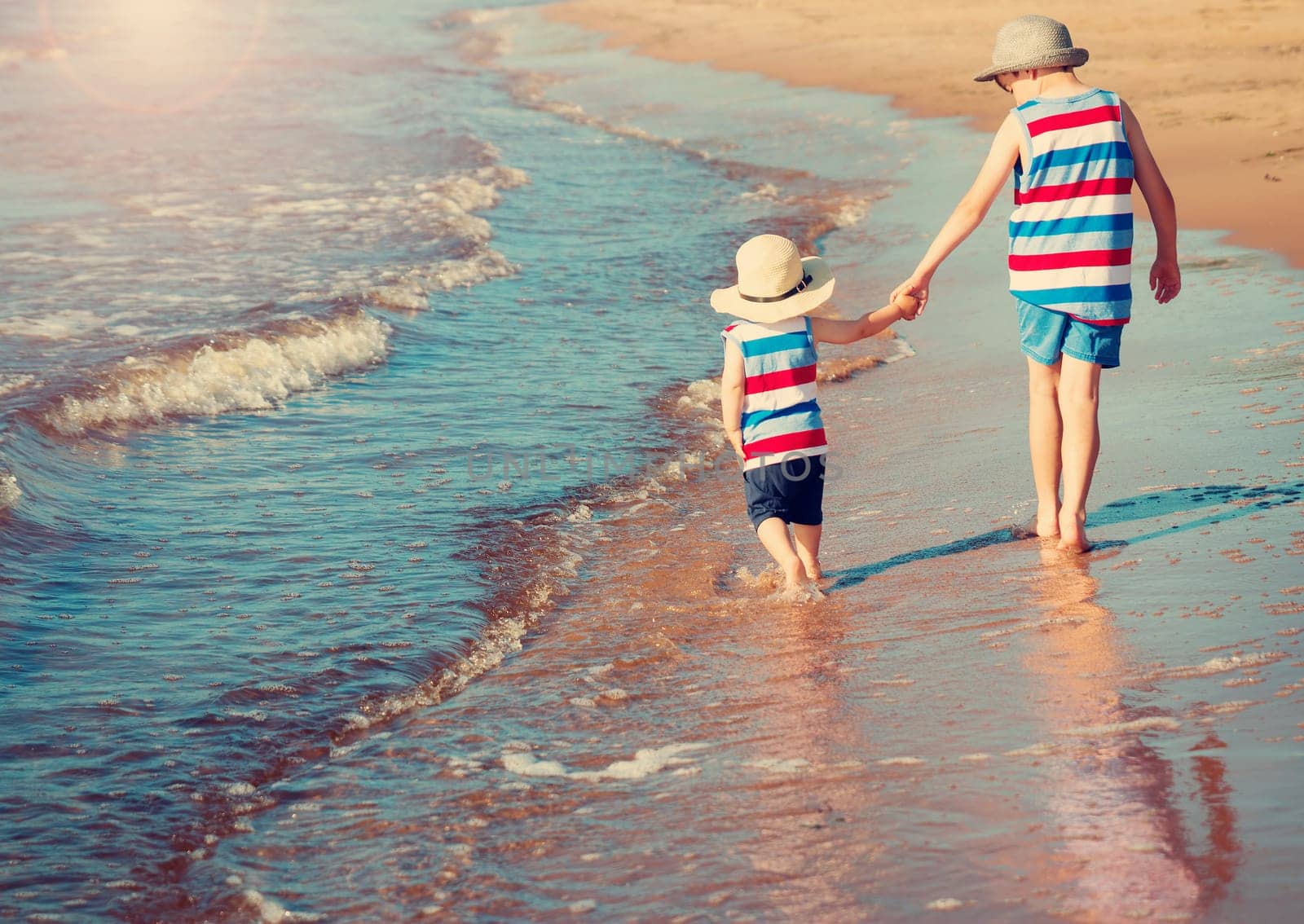 Two brothers barefoot are walking on the beach in water. Boys are on vacation in summer at the sea