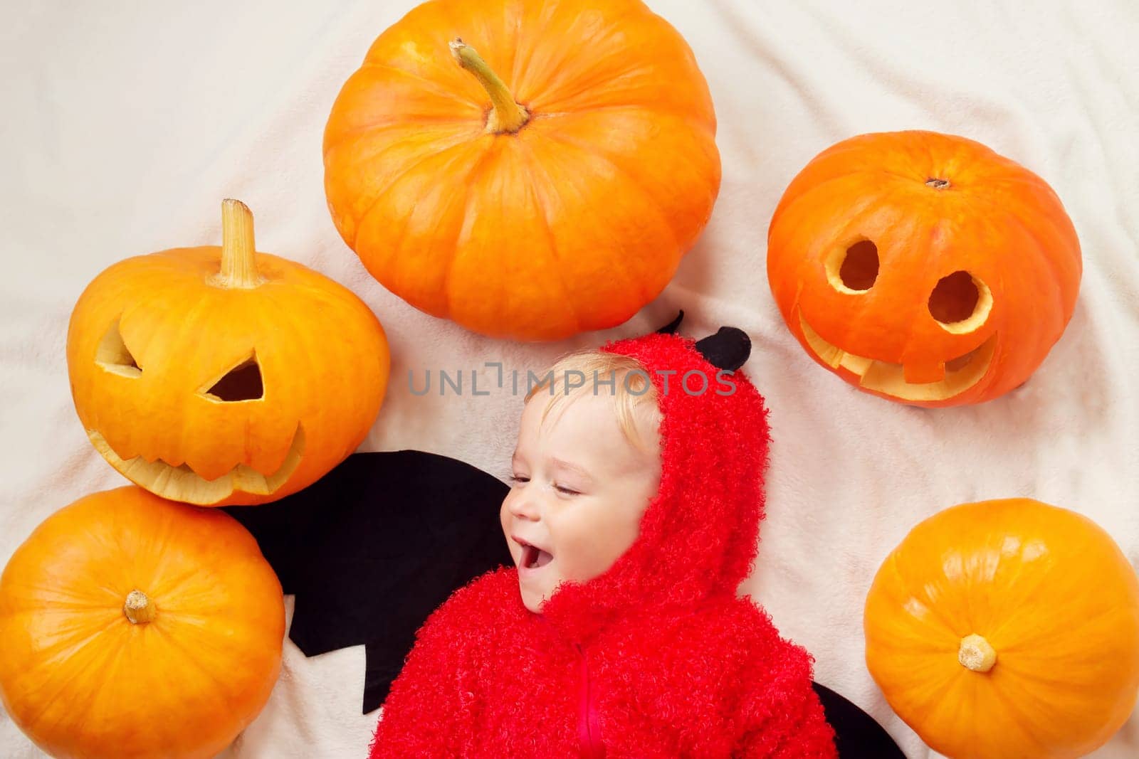 Baby in devil costume with pumpkins. Child in halloween outfit lying on soft white blanket