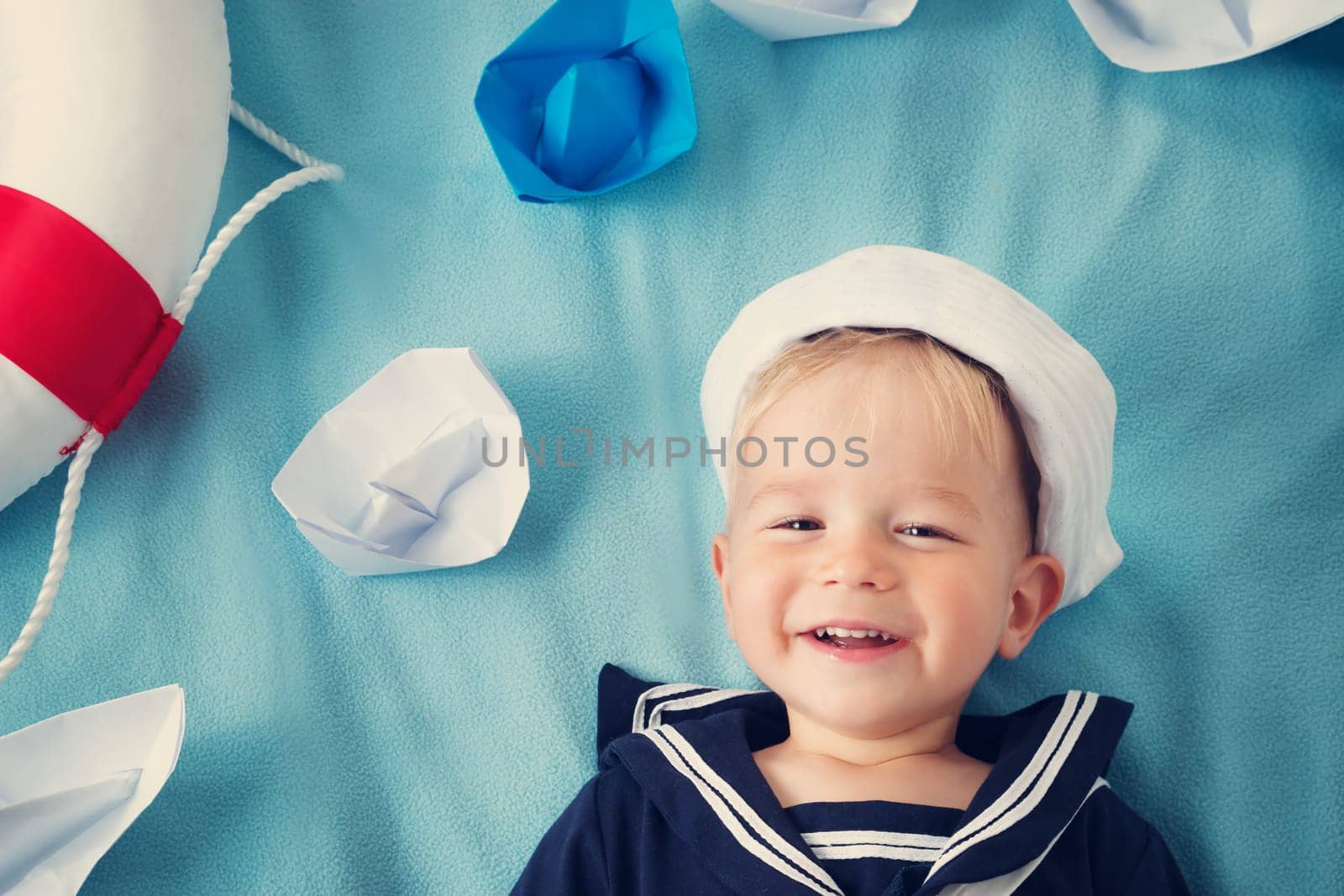 two years old boy playing in sailor hat. Child with paper ships on blue background