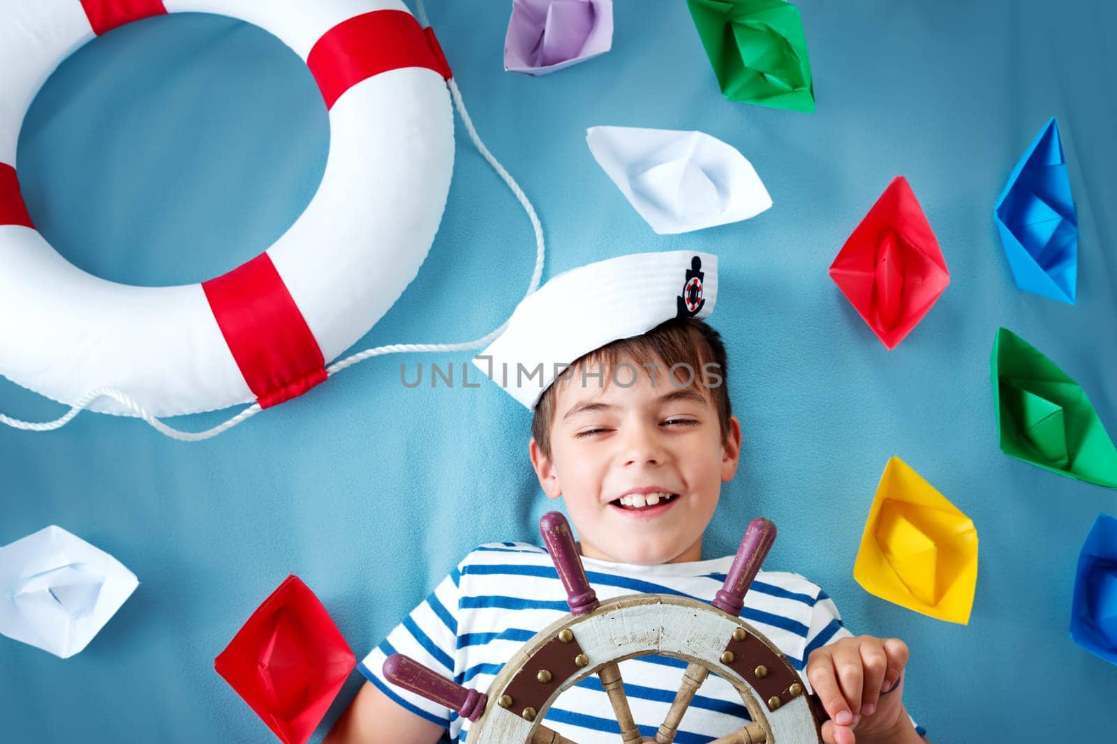 seven years old boy playing in sailor hat. Child with a steering wheel and paper ships on blue background