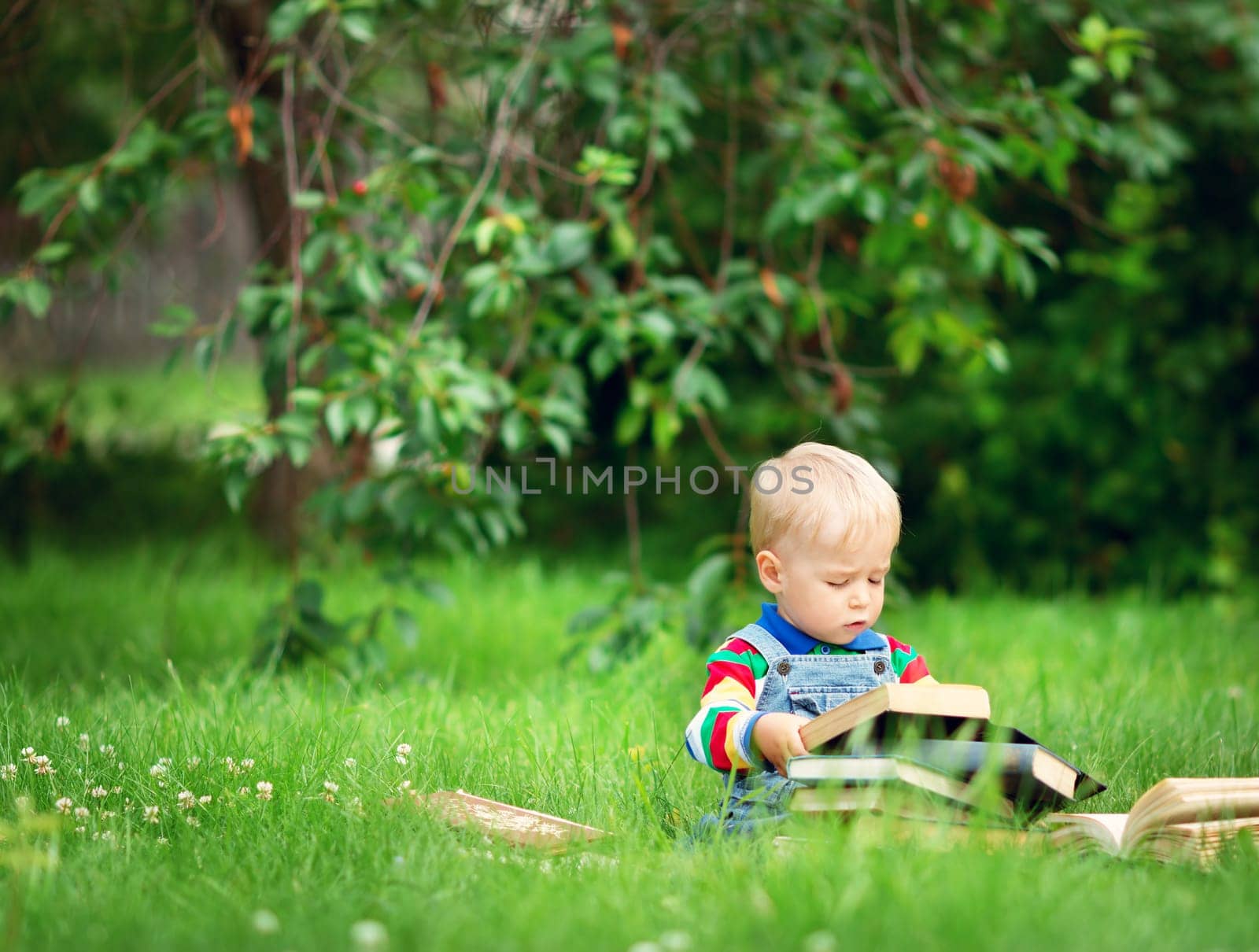 one year old baby reading a book sitting on the grass in garden