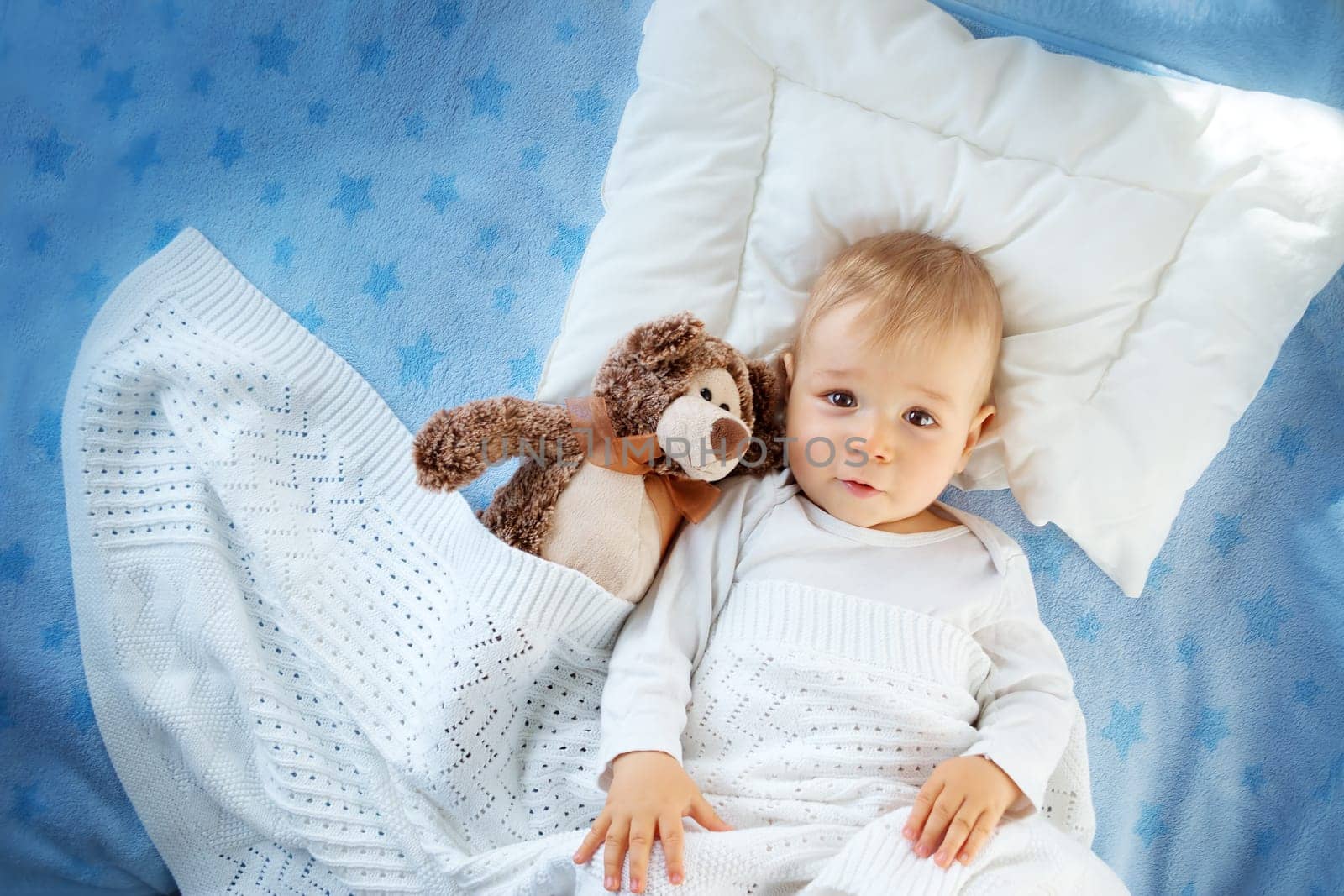 One year old baby lying in bed with a plush teddy bear. Child sleeping on soft blue blanket with stars