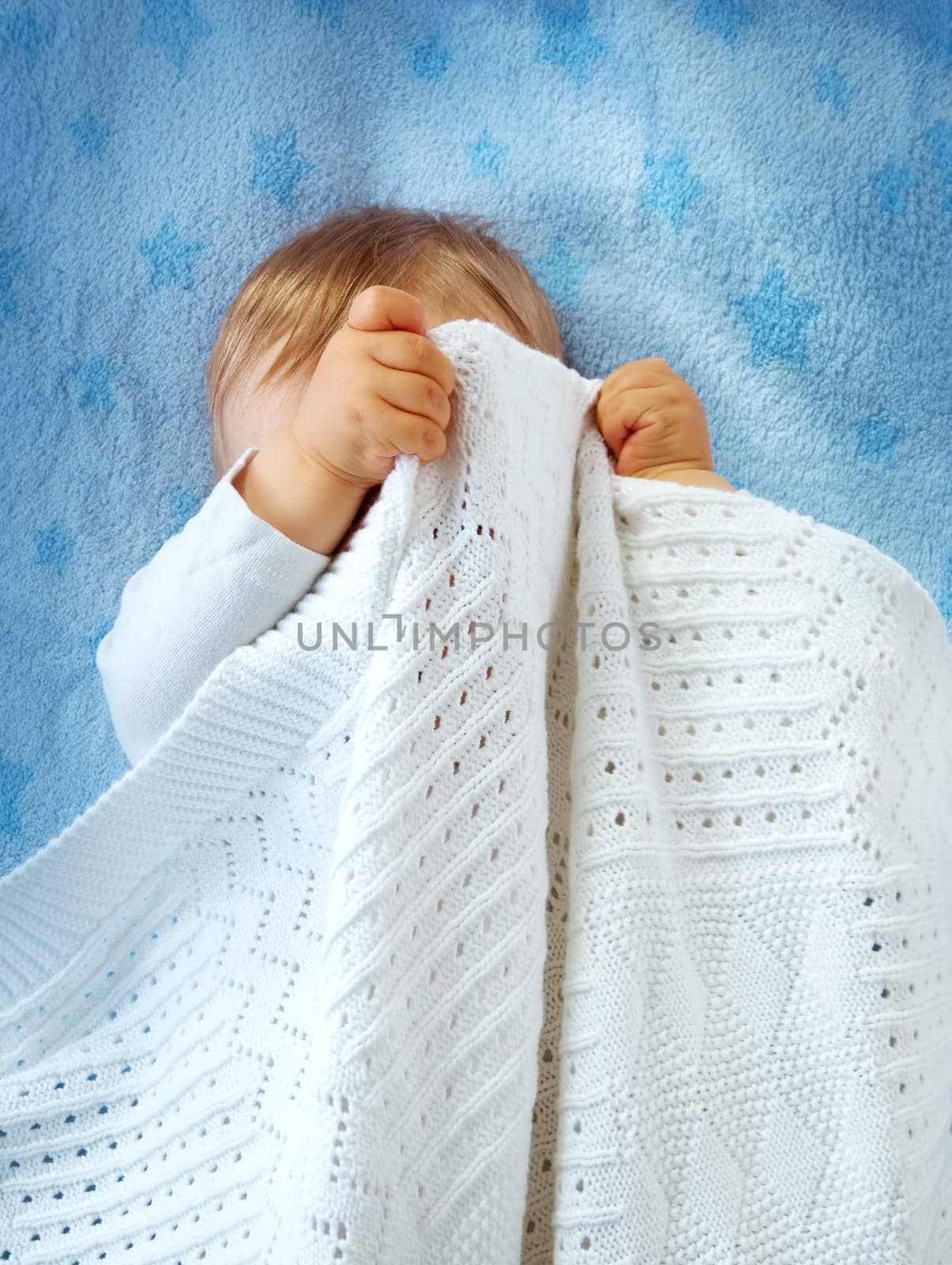 One year old baby lying on blue towel with stars. Child hiding under blanket