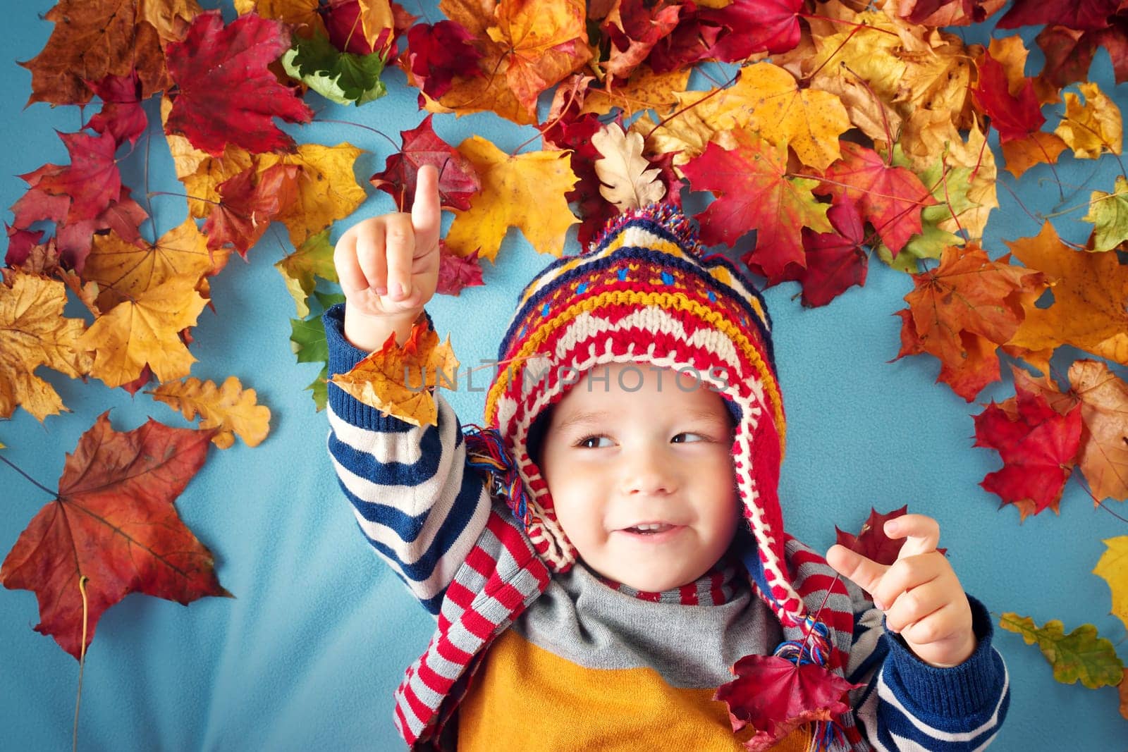 two years old boy dreaming in autumn. Child in hat and scarf playing with maple leaves on blue background