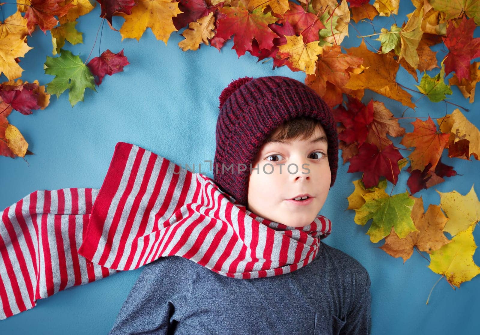 seven years old boy dreaming in autumn. Child in hat and scarf and maple leaves on blue background