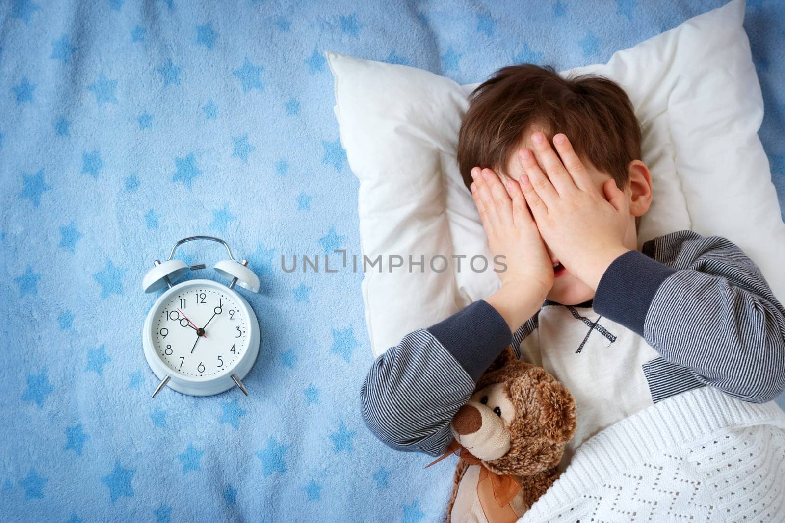 six years old child sleeping in bed on pillow with alarm clock and a teddy bear