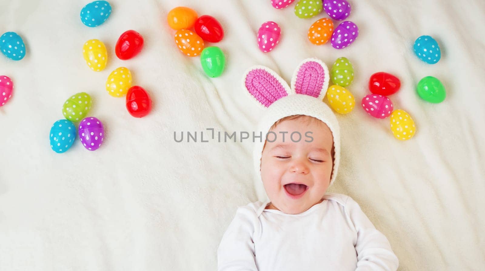 Baby boy in bunny hat lying on green blanket with multicolored easter eggs