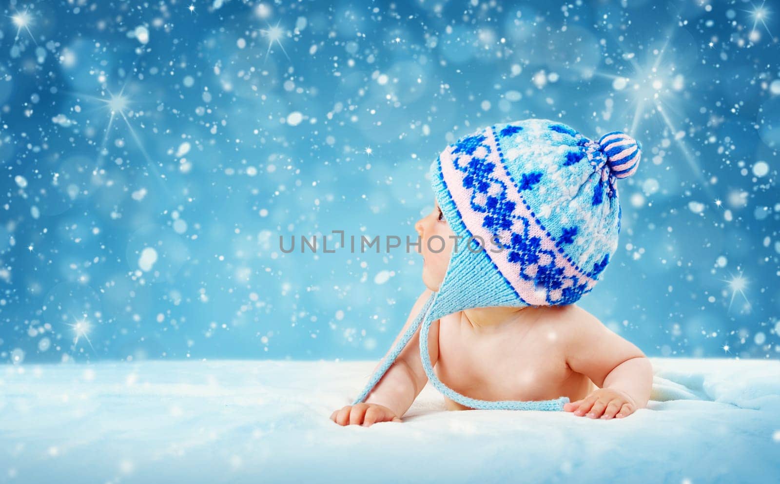 Eight month old baby in knitted winter hat lying on soft blanket with blue background and falling snow