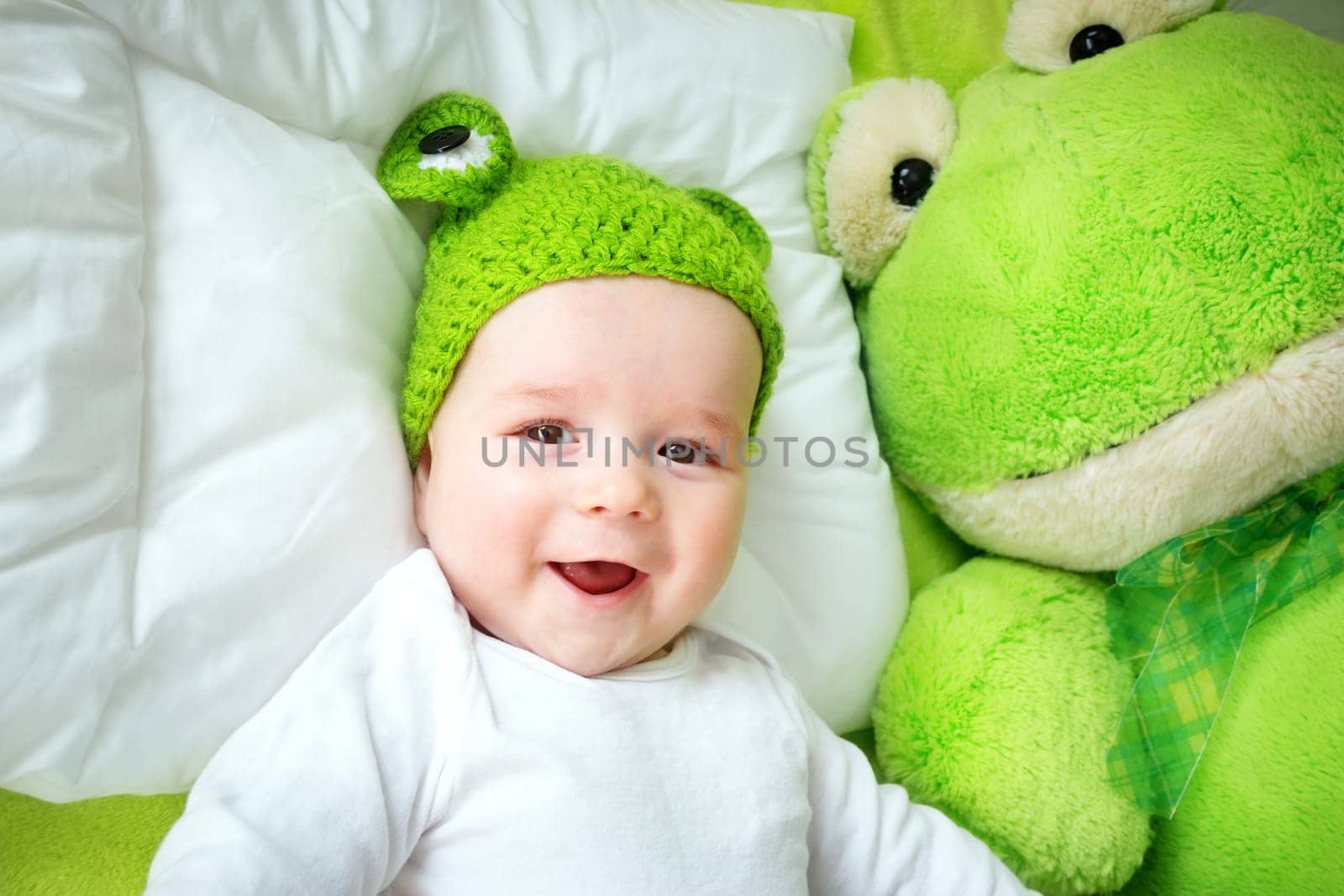 little boy in knitted animal hat lying on soft green blanket with frog softtoy. Cute 5 month old baby on the pillow in bed