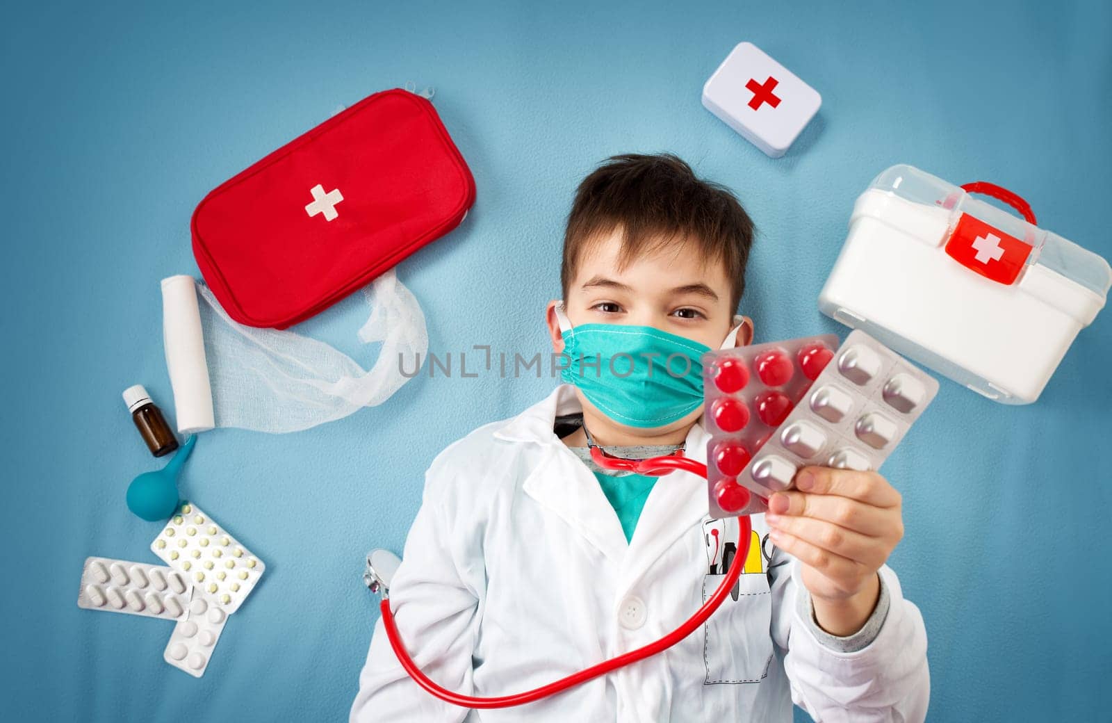 Child playing doctor in costume and mask. Boy holding pills with medical accessories around.