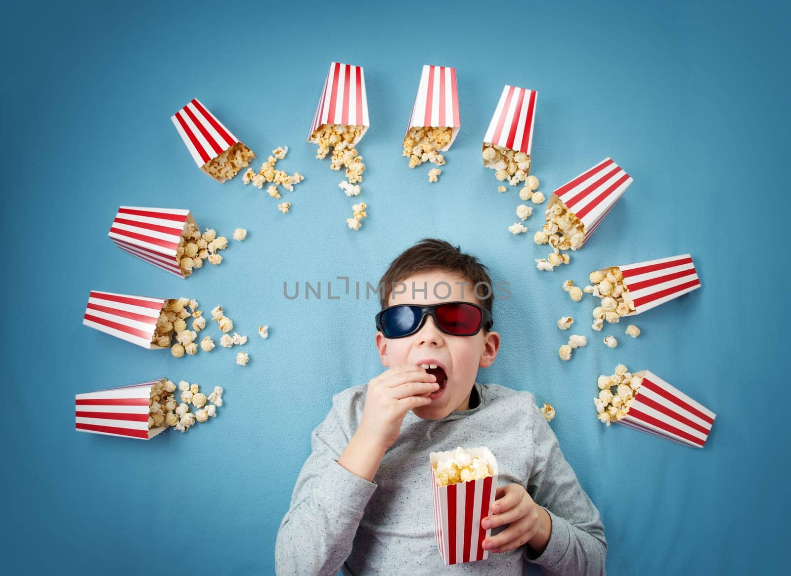 Child lying on blue background in 3d glasses and watching tv. Boy eating popcorn