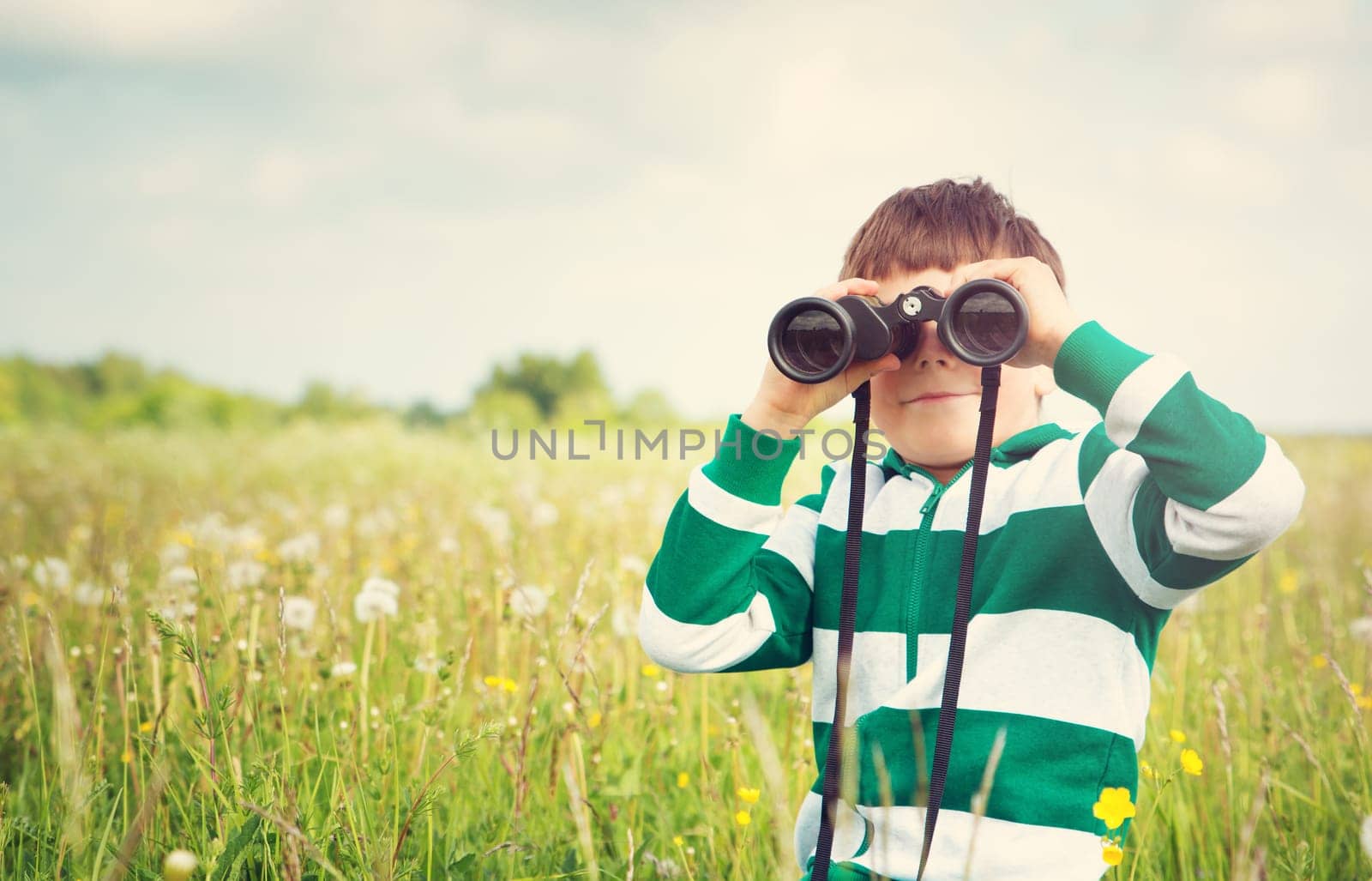 Child wathing in benoculars on the field with dandelions. Boy playing on the meadow in summer