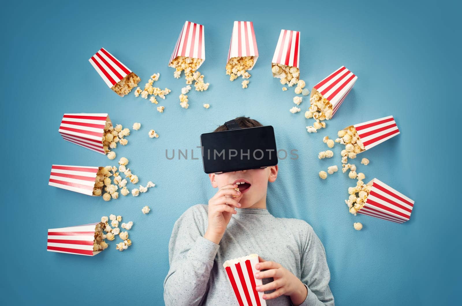 Child lying on blue background in Virtual reality glasses and watching film. Boy eating popcorn