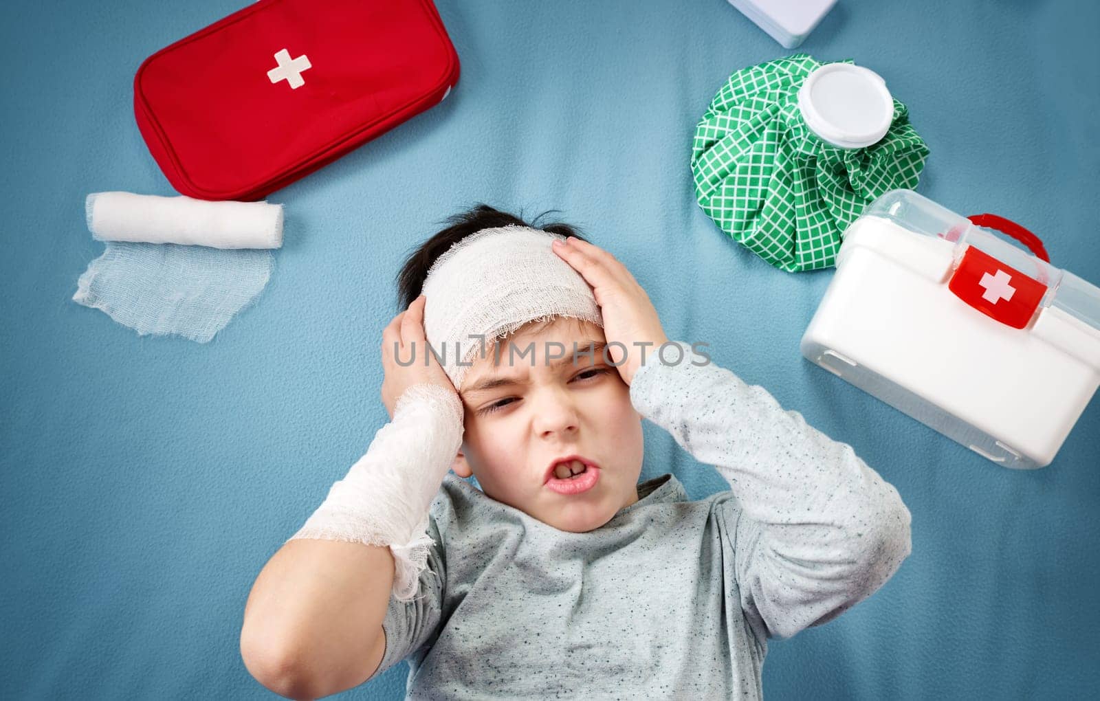 Sad child in bandages lying in bed. Wounded boy on blue background with first aid accessories.