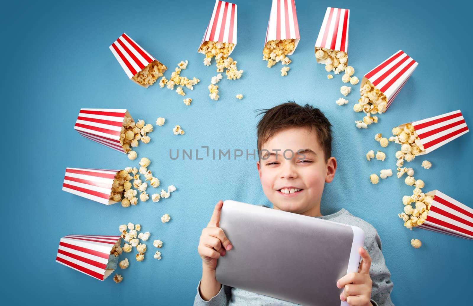 Child lying on blue background with tablet and watching movie. Boy with popcorn
