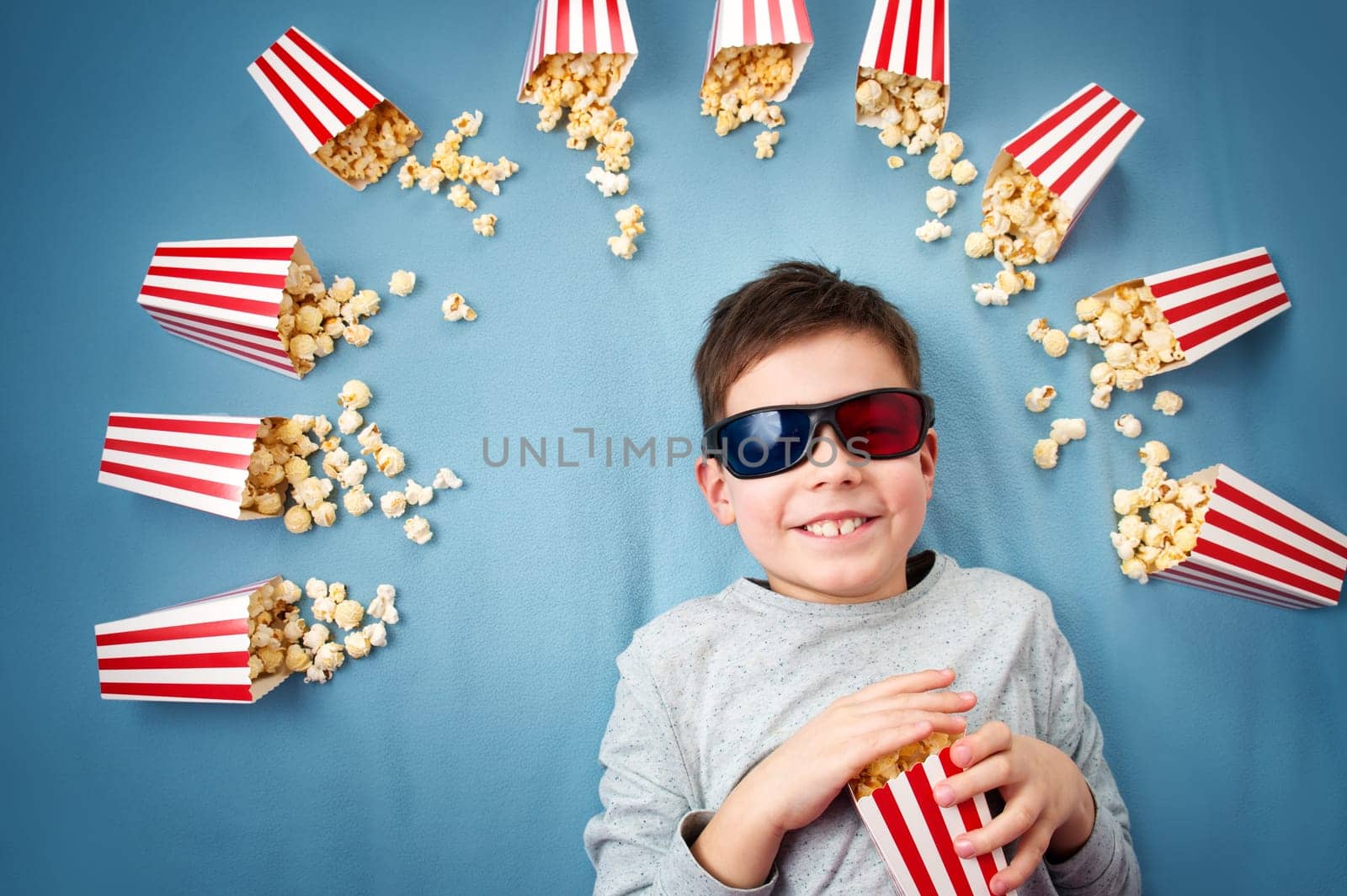 Child lying on blue background in 3d glasses and watching tv. Boy eating popcorn