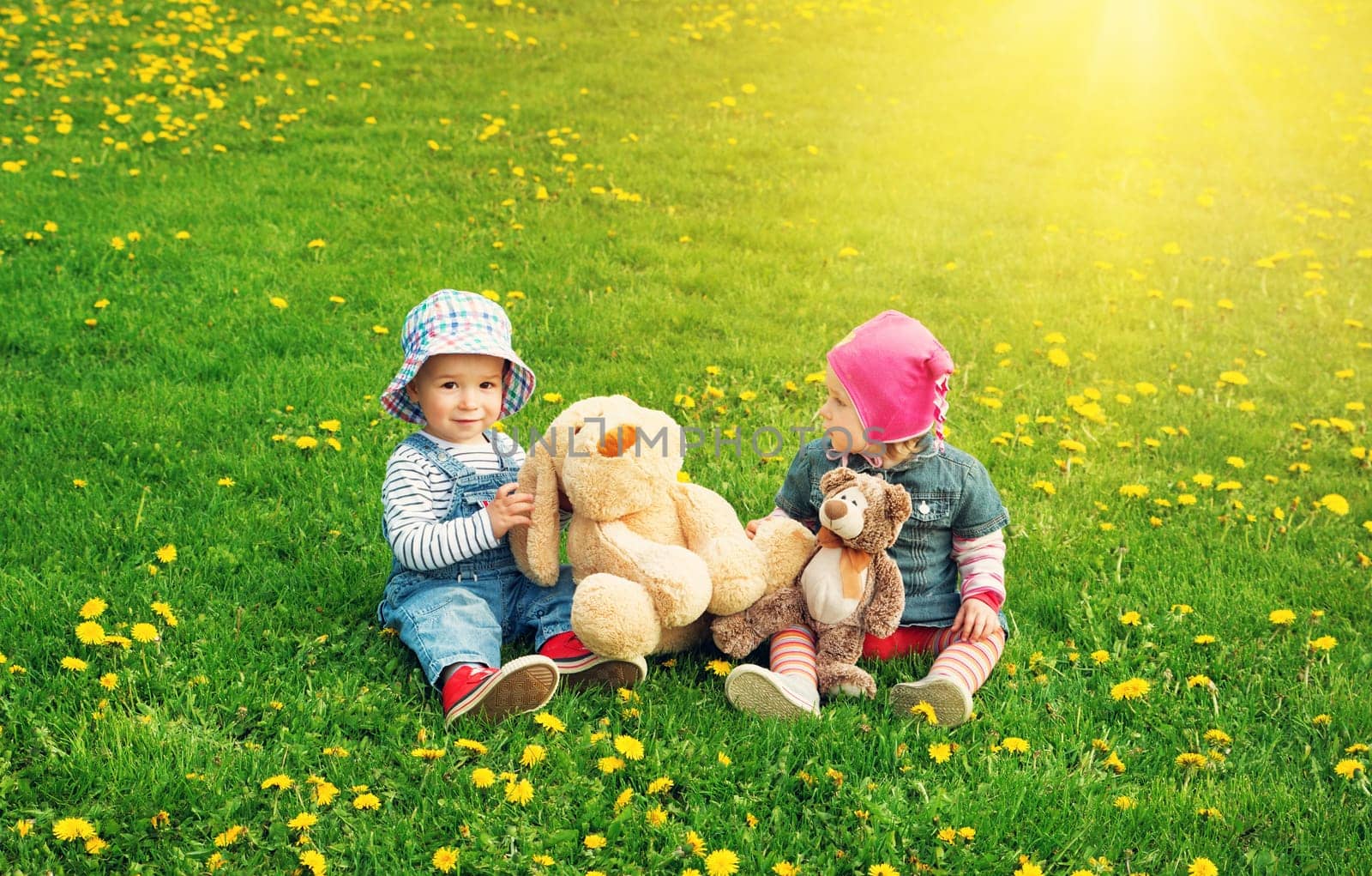 Little boy and girl in hats sitting on the field with soft toys in summer. Two cute one year old babies on the grass with dandelions