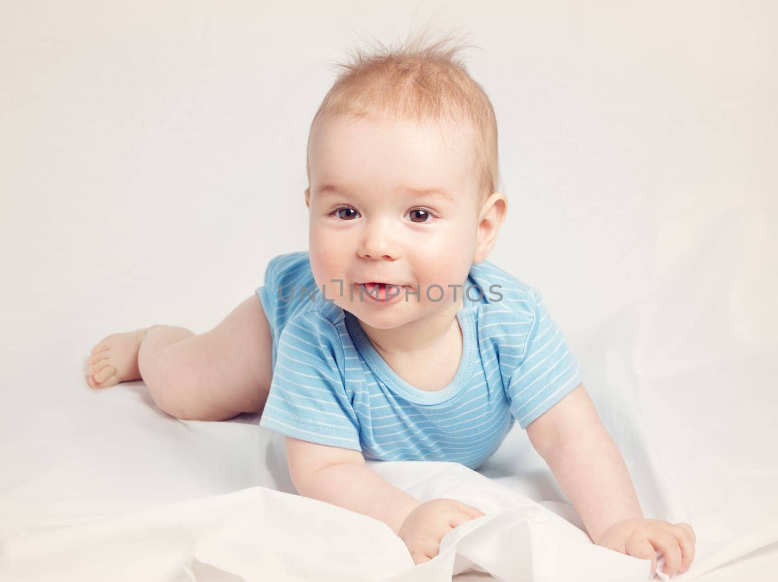 Eight month old baby lying in the bed on white sheet. Cute boy showing tongue
