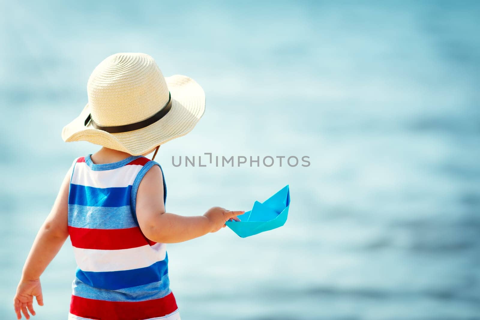one year old boy playing at the beach in straw hat. Child with a paper ship at sea