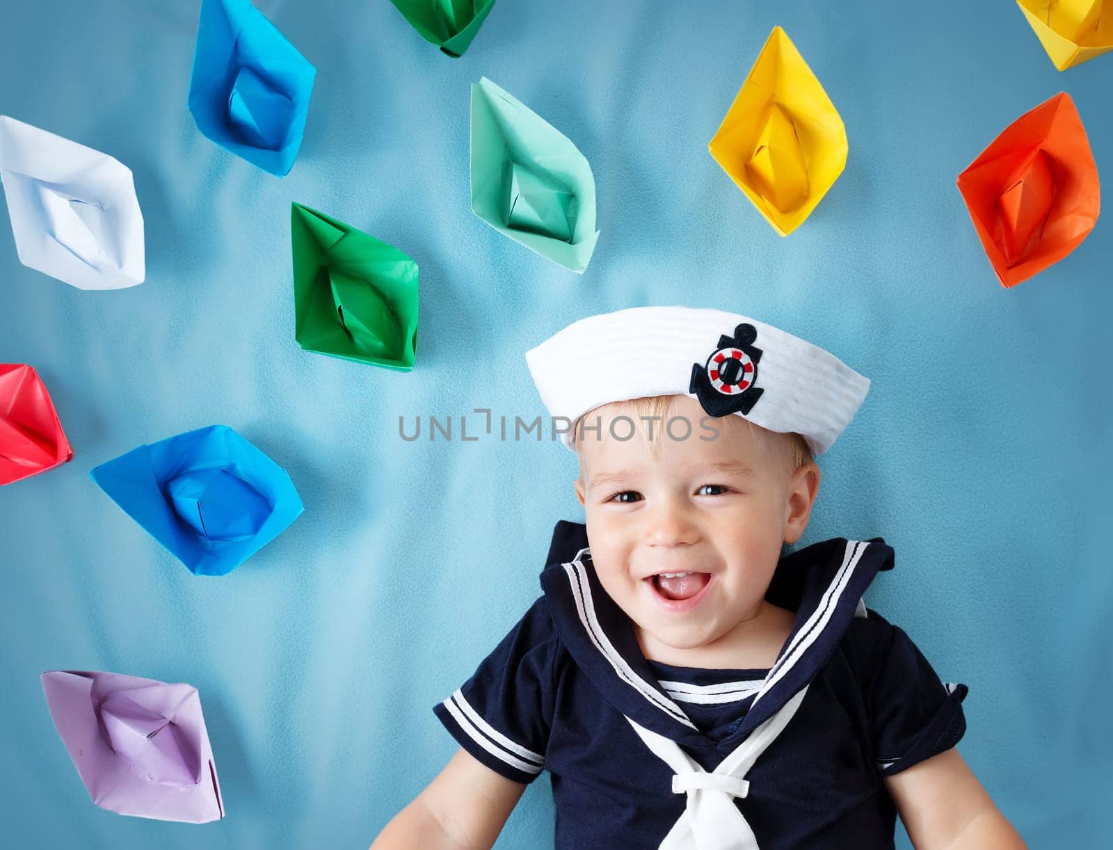 two years old boy playing in sailor hat. Child with paper ships on blue background