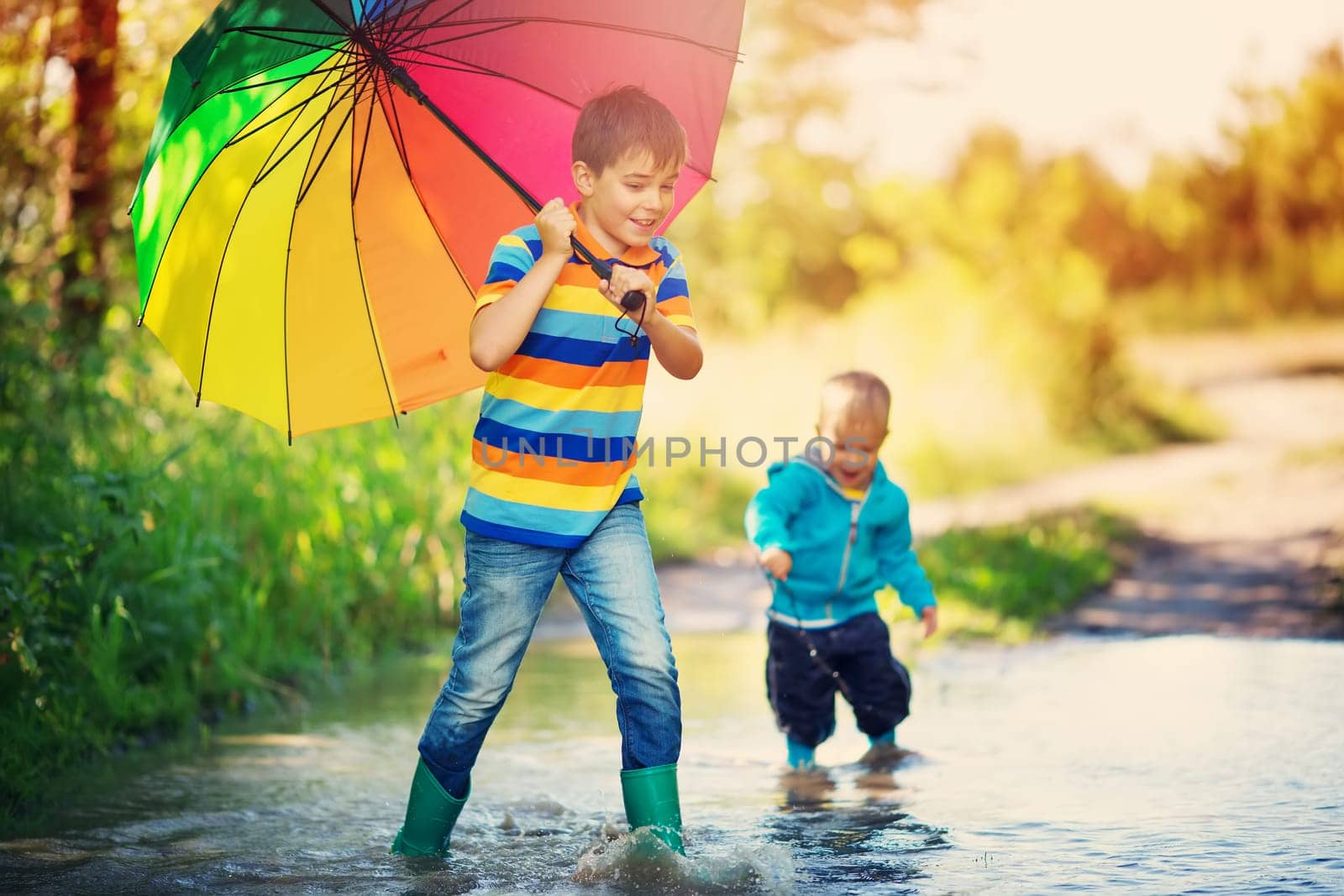 Children walking in wellies in puddle on rainy weather. Boy holding colourful umbrella under rain in summer