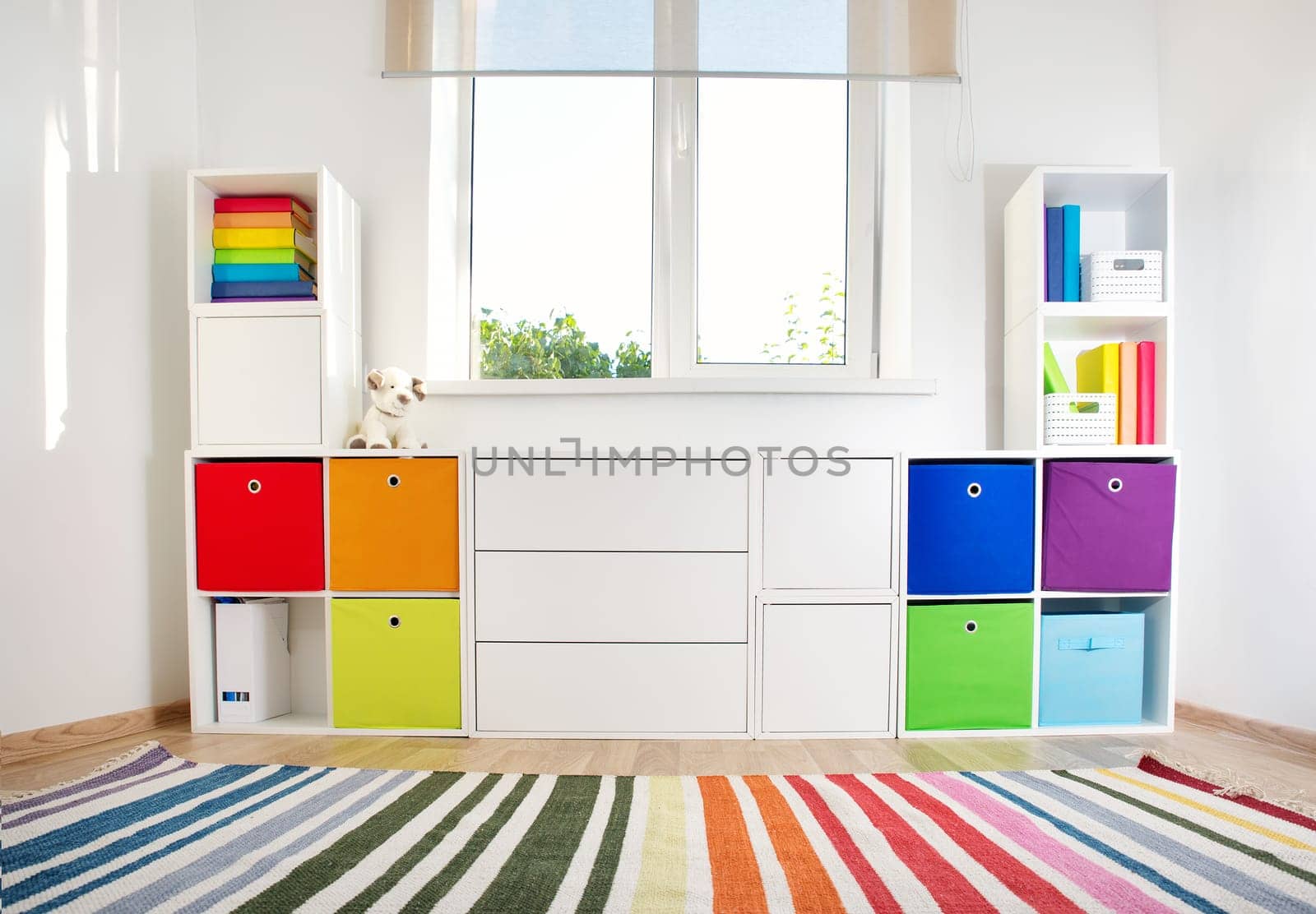 Colourful children rooom with white walls and furniture. Rainbow carpet at home interior with a window