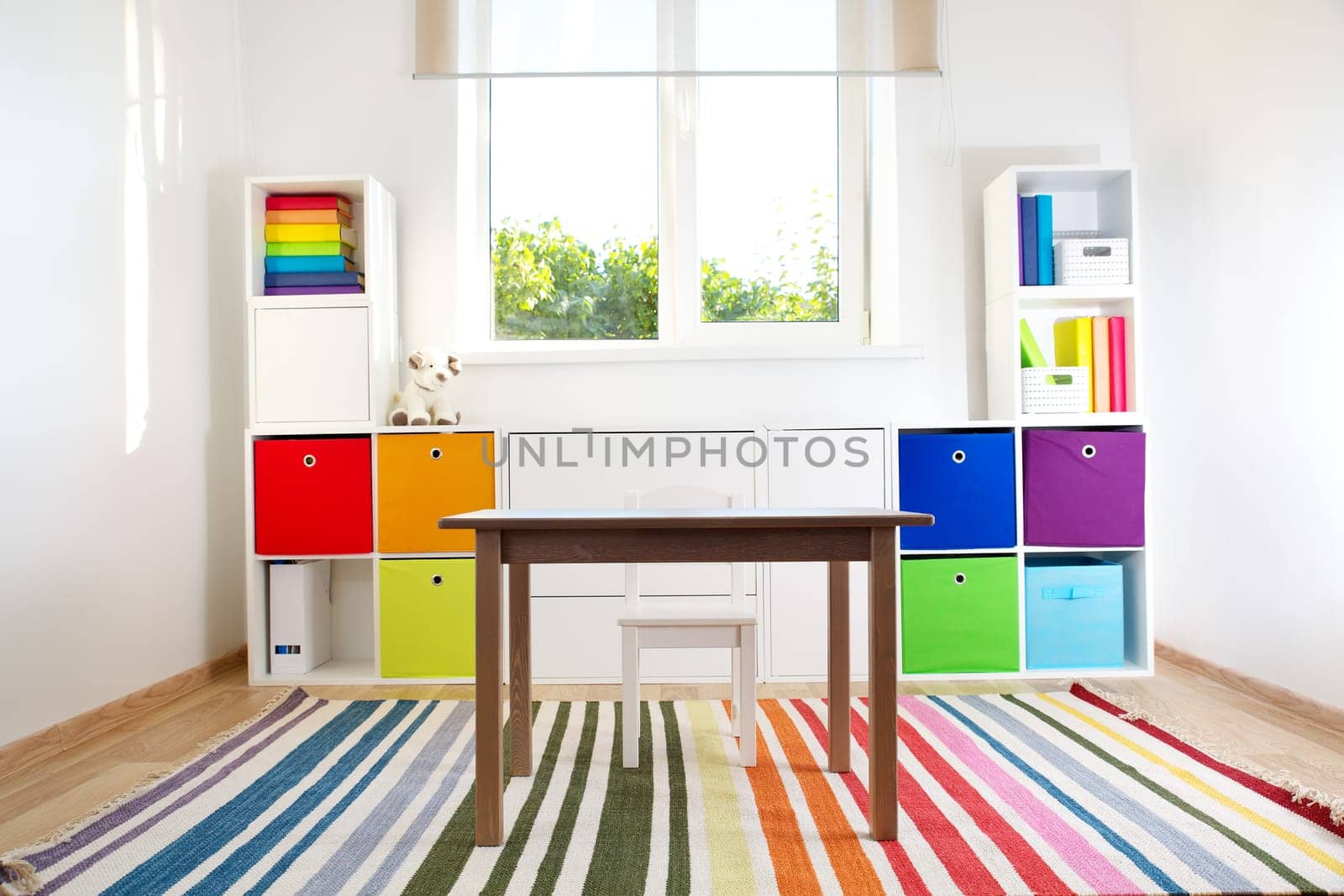 Colourful children rooom with white walls and furniture. Rainbow carpet at home interior with a window