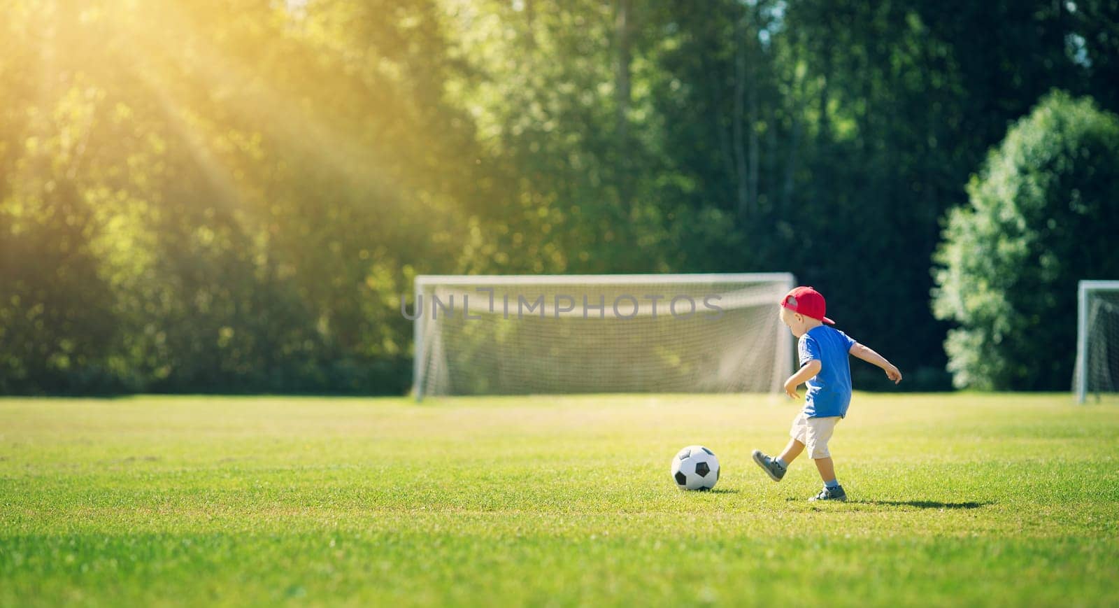 Little boy playting football on the field with gates. Active child in summer with ball