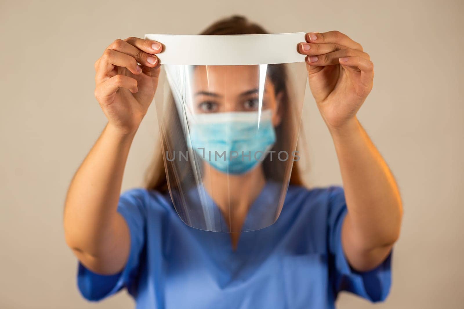 Female nurse in blue shirt holding protective face shield in front of her. Concept of medical safety during pandemic of covid-19. Young woman with brown hair using protection against viruses.