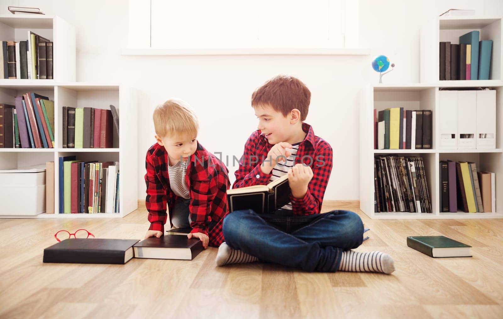 Three years old child and his older brother sitting among books at home. Curious boys reading in white room