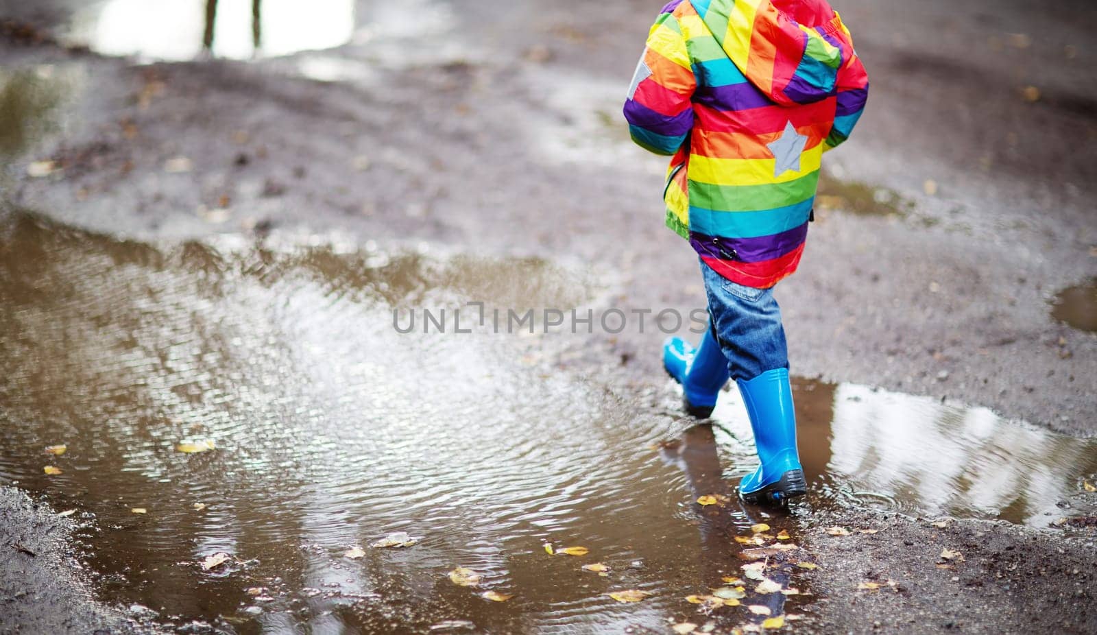 Child walking in wellies and jumping in puddle on rainy weather. Boy under rain in summer outdoors