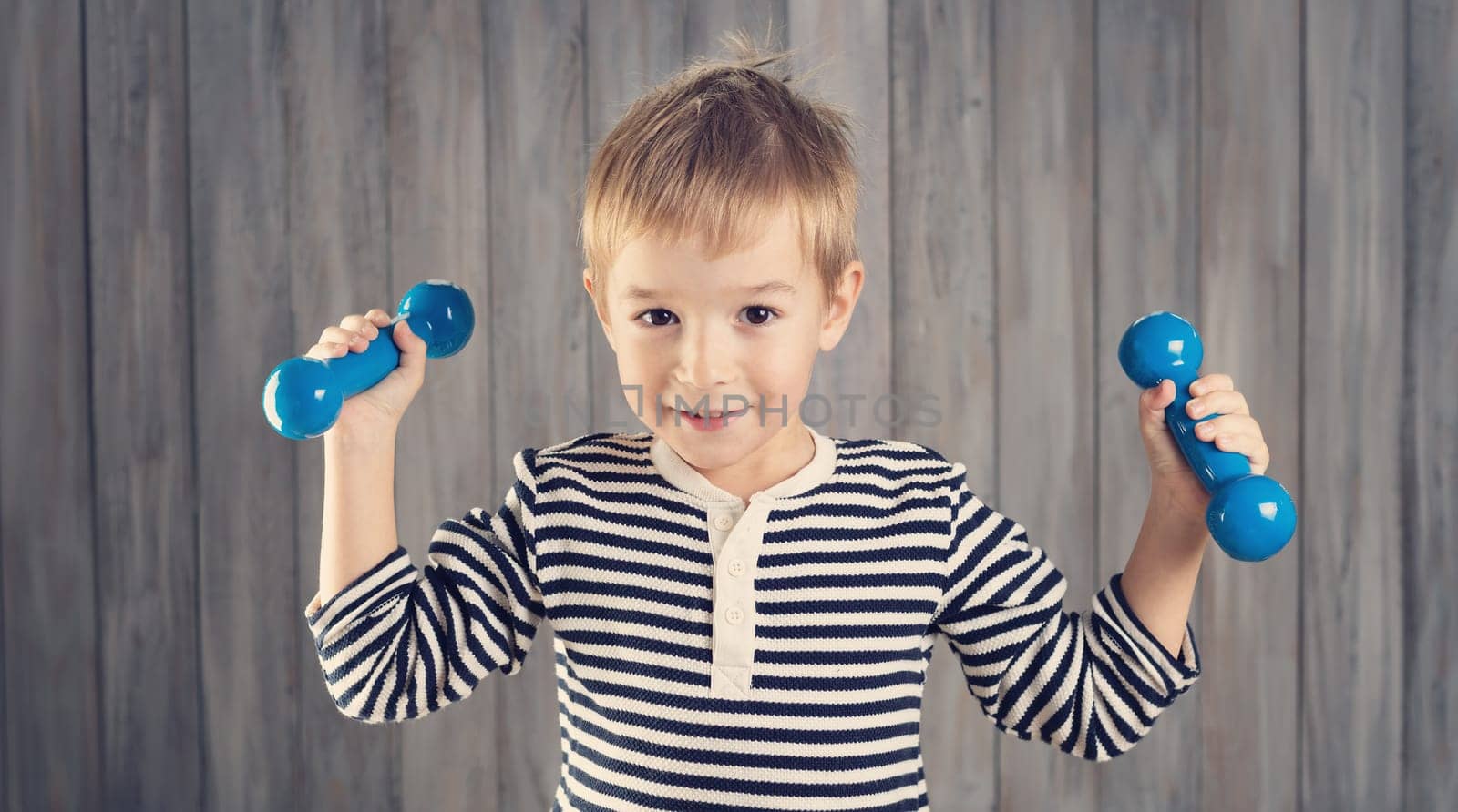 Child holding dumbbells indoors on wooden background. Happy active boy playing fitness. Healthy lifestyle