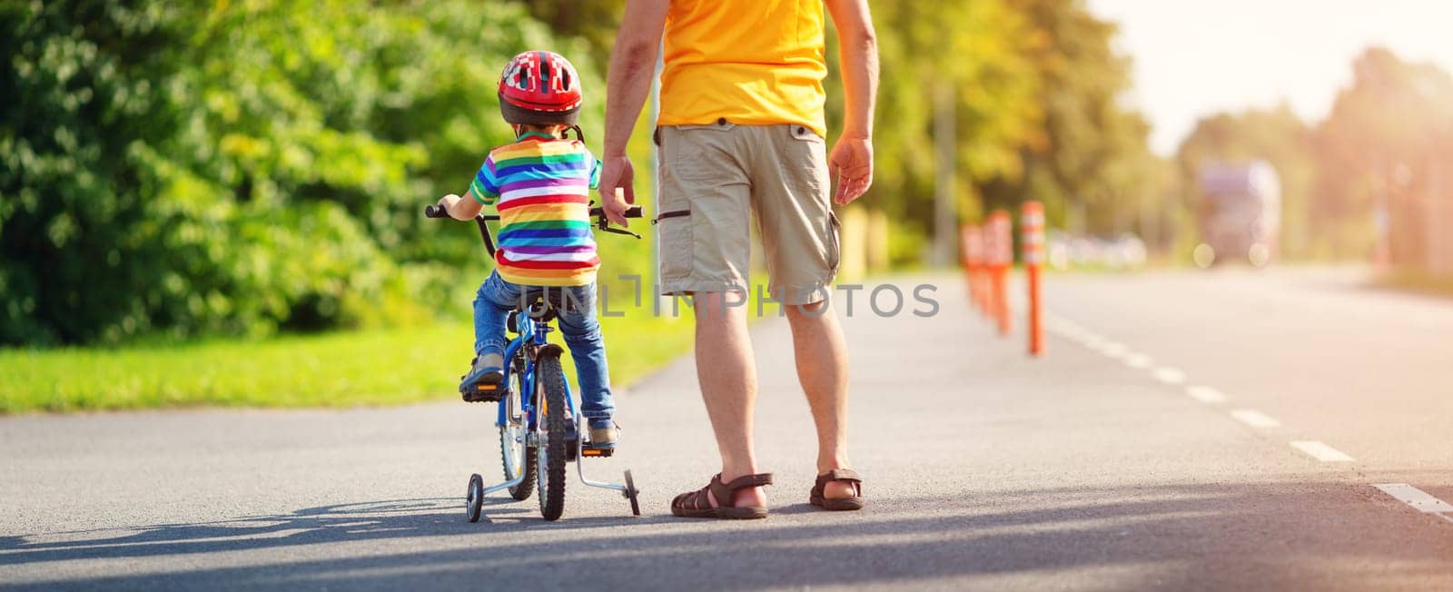 a child on a bicycle in helmet with father on asphalt road. Little boy learning to ride on bike with dad outdoors on sunny summer day.