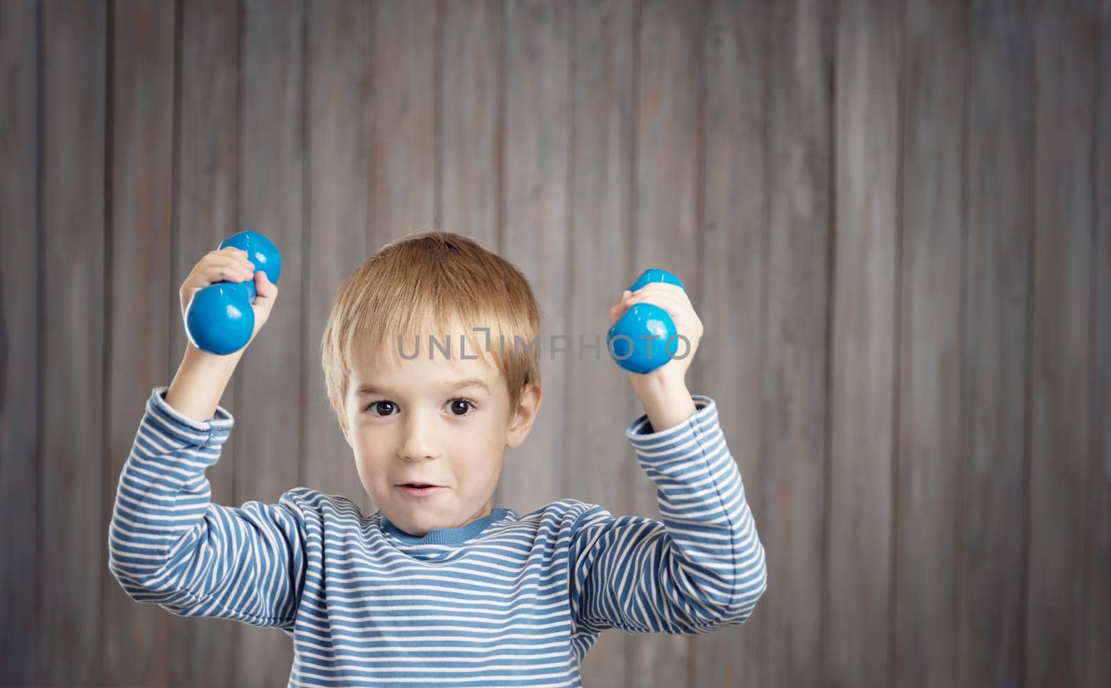 Child holding dumbbells indoors on wooden background. Happy active boy playing fitness. Healthy lifestyle