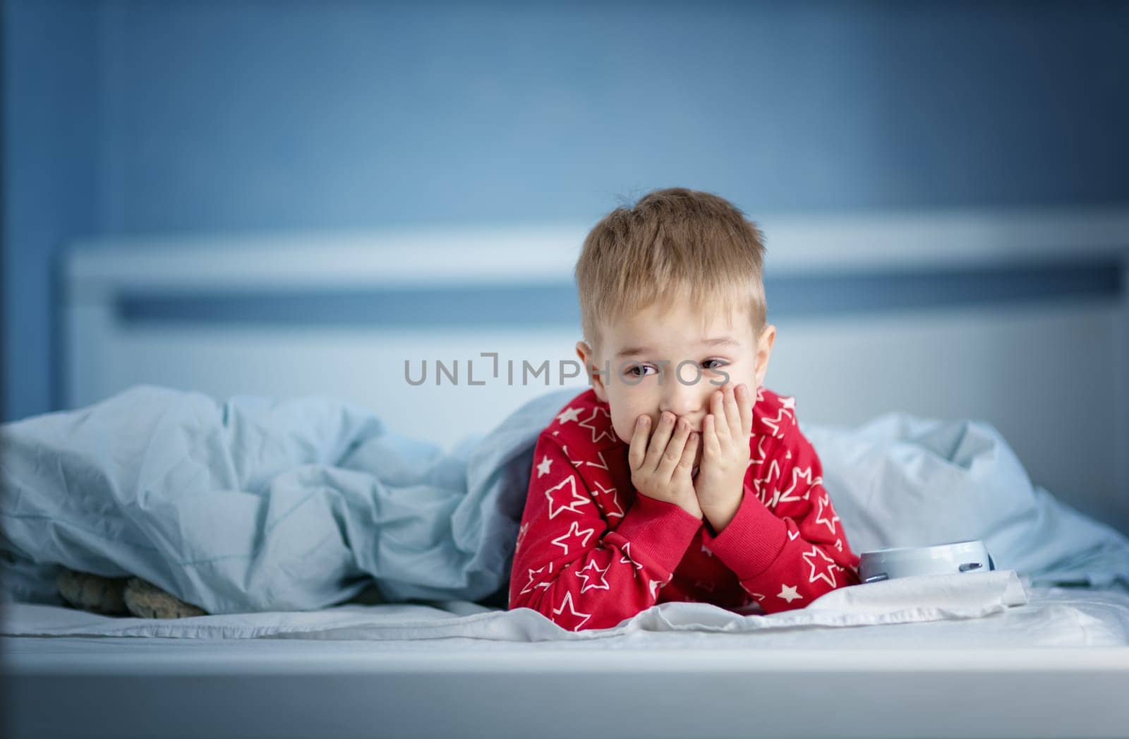 Sleepy boy lying in bed with blue beddings. Tired child in bedroom. Little kid lying awake in red pajamas with toothache