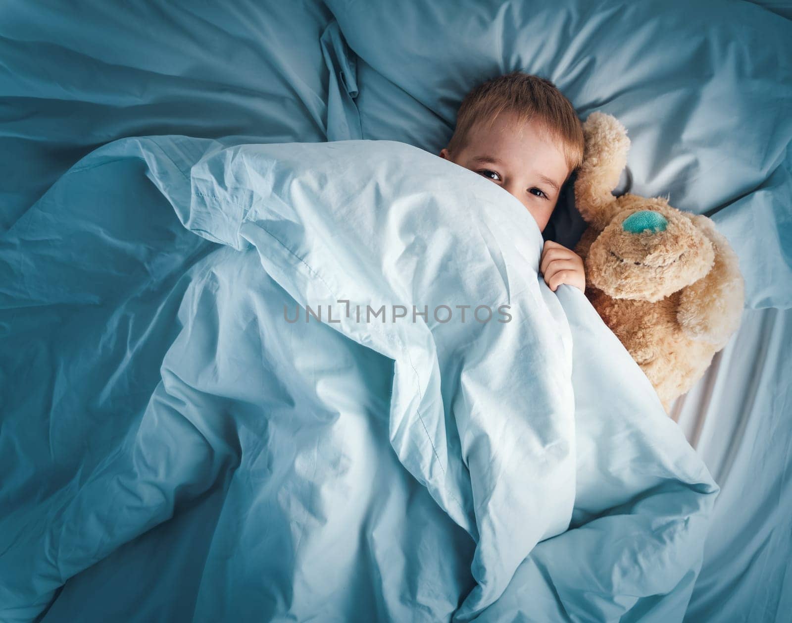 Sleepy boy lying in bed with white beddings. Tired child in bedroom sleeping. Little kid lying asleep with toy