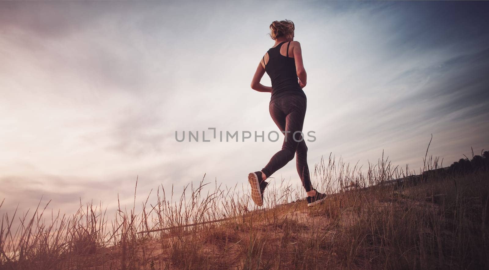 Young woman running on the field near seaside at sunset. Active person outdoors at the dusk in summer