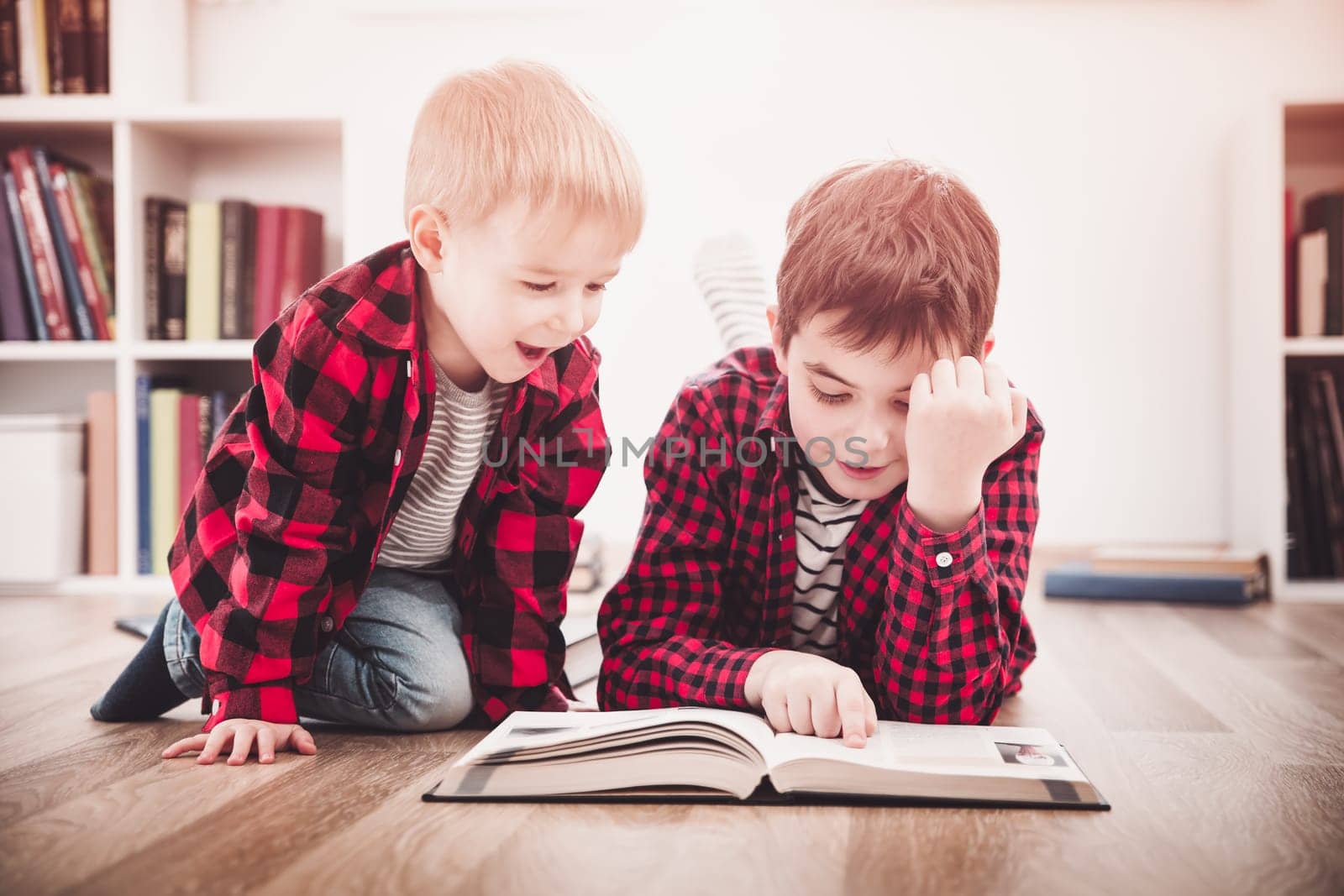 Three years old child and his older brother sitting among books at home. Curious boys reading in white room