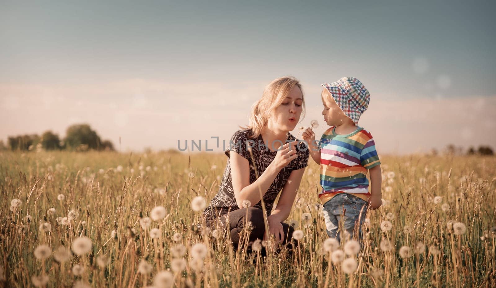 Young woman with a boy blowing dandelions. Mother and son having fun on the field