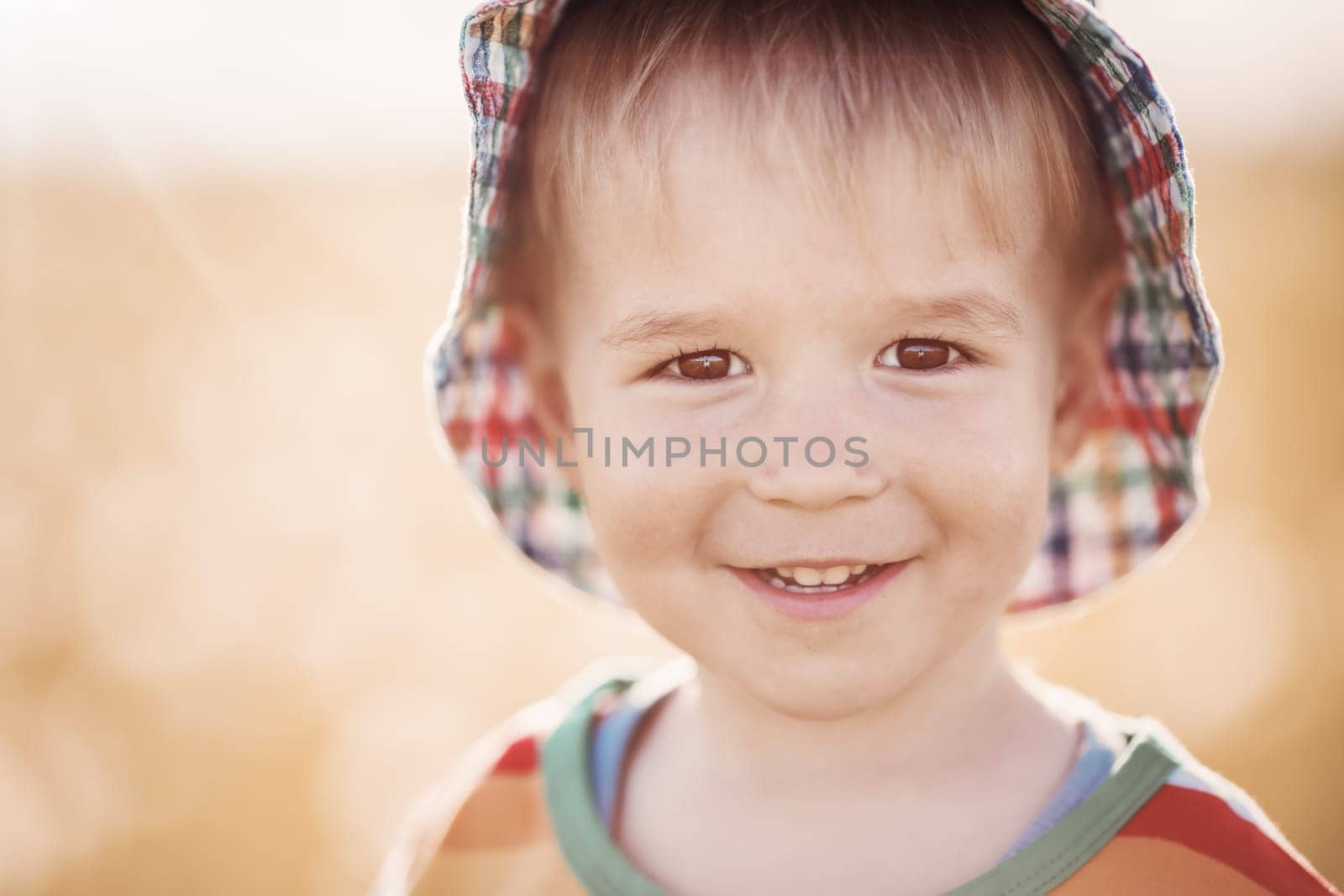 Happy baby boy standing in grass on the fieald with dandelions at sunny summer evening. child outdoors in hat