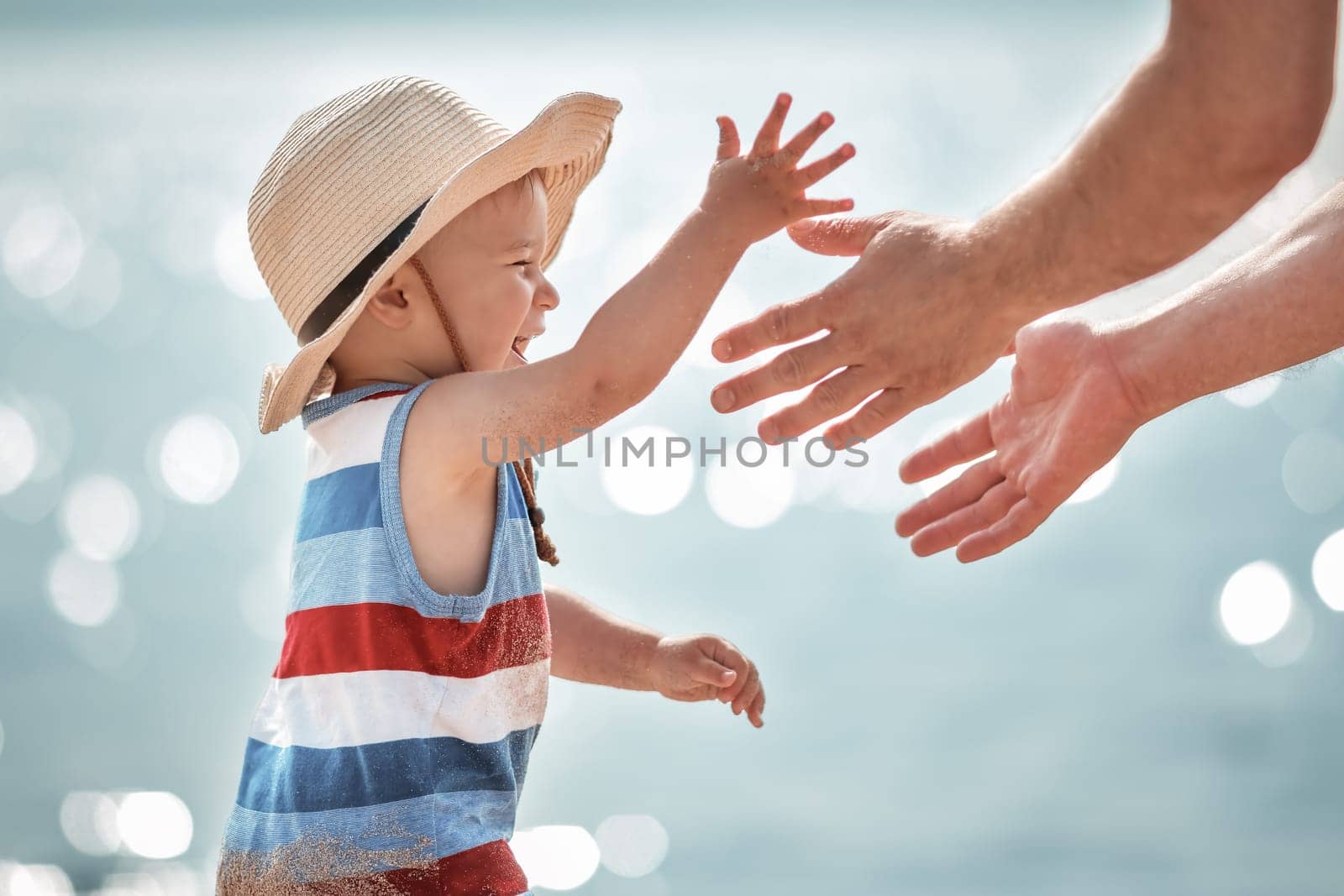 one year old boy playing with father at the beach in straw hat. Child on family vacations at sea. Mans hands and a baby