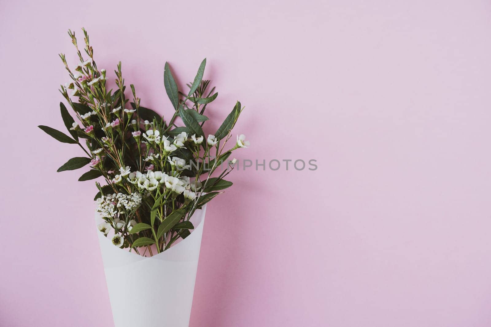 top view closeup of beautiful bouquet of delicate small white flowers with green leaves in paus tracing paper cone on pink background copyspace available