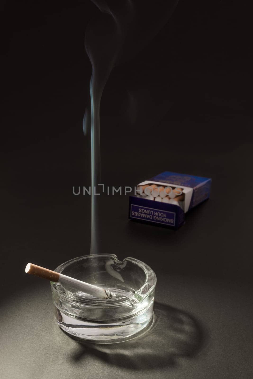 A lid cigarette lies burning in an ashtray, with the smoke floating up in to the air, with an open packet of cigarettes in the background.