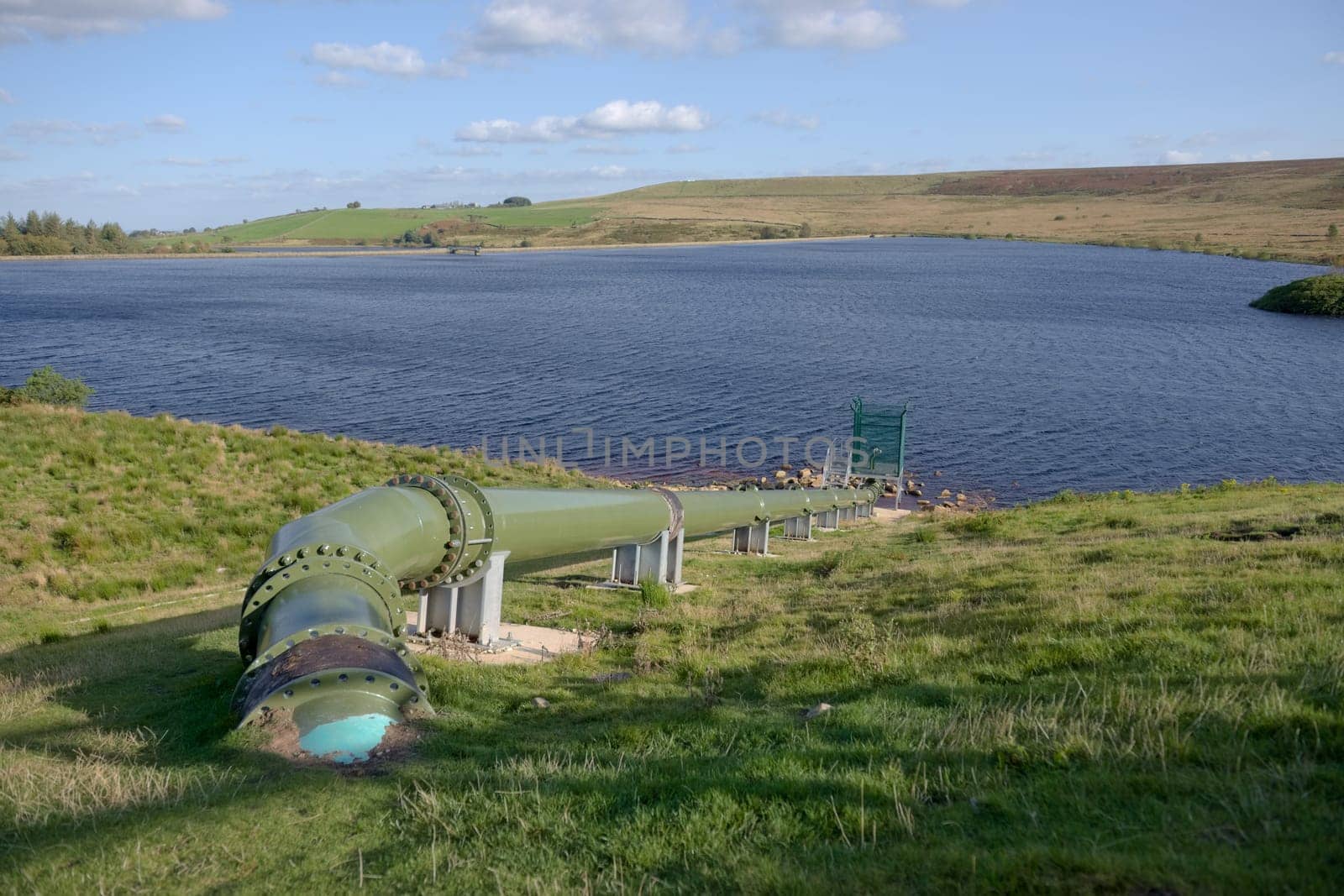 Warm sunlight shines on a shiny green pipe feeding water into a deep blue rural reservoir. Cloudy sky background. Modern industrial hydro equipment.