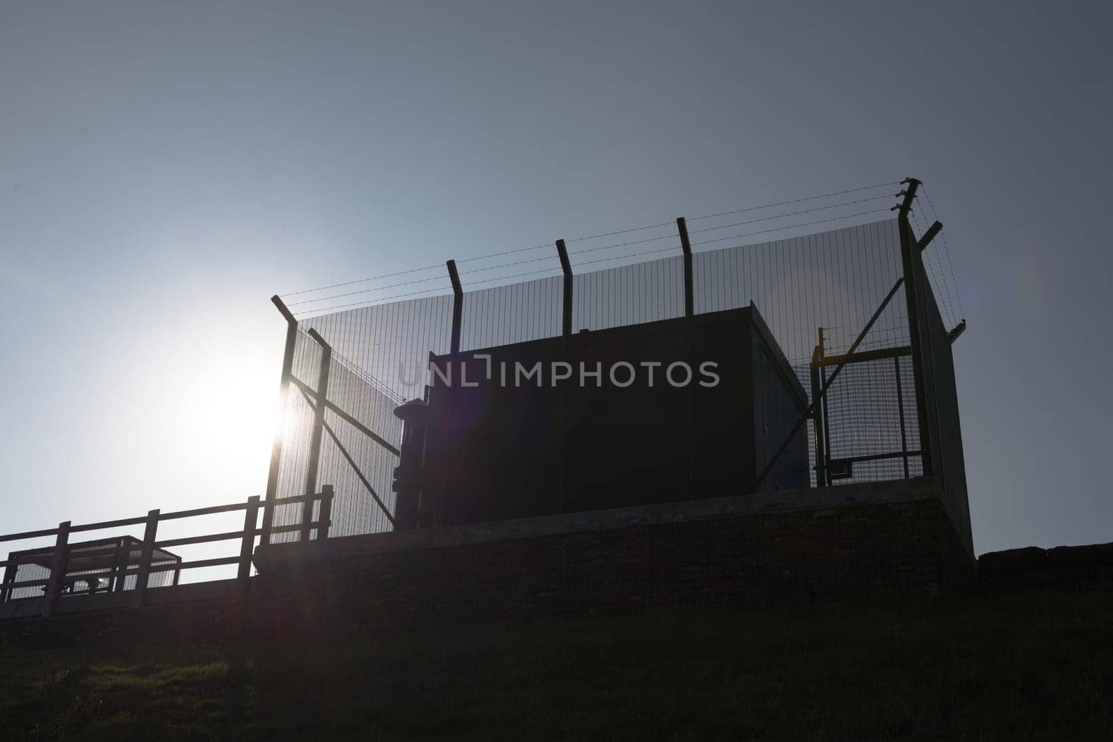 Tall barbed wire anti climb fence encases large square metal industrial water management equipment. Looking up, plain blue sky and bright sun behind