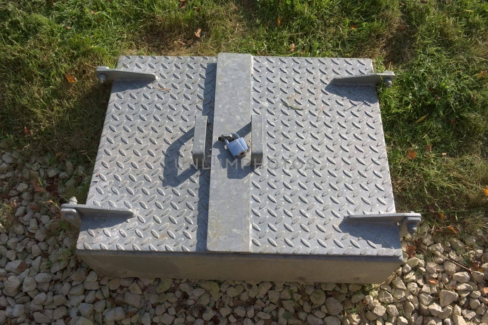 Modern secure padlocked steel service hatch in rural sunlight. Manhole cover juxtaposed against natural surroundings. Shadowed checkered steel texture